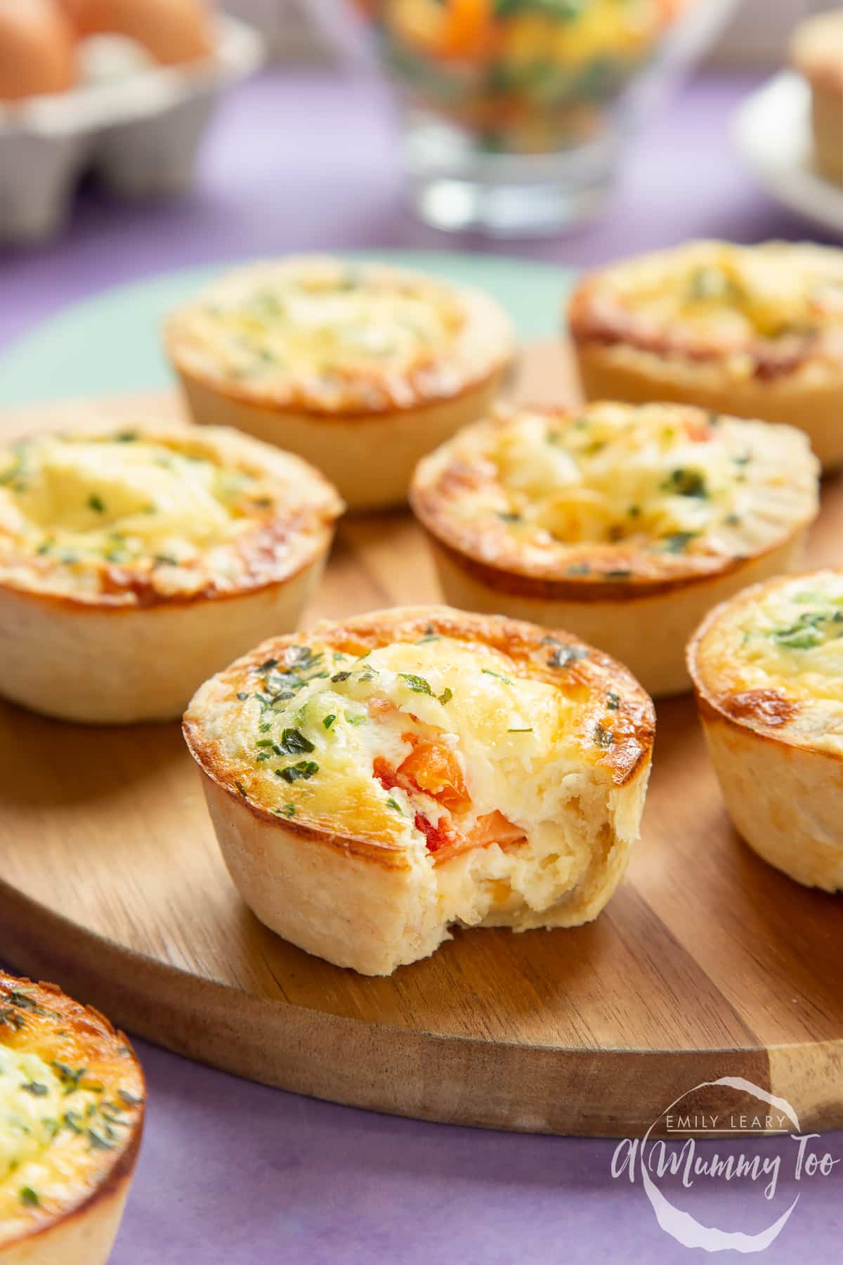 A mini vegetable quiche with a bite out of it, revealing the vegetables inside. More are arranged on a wooden board in the background.