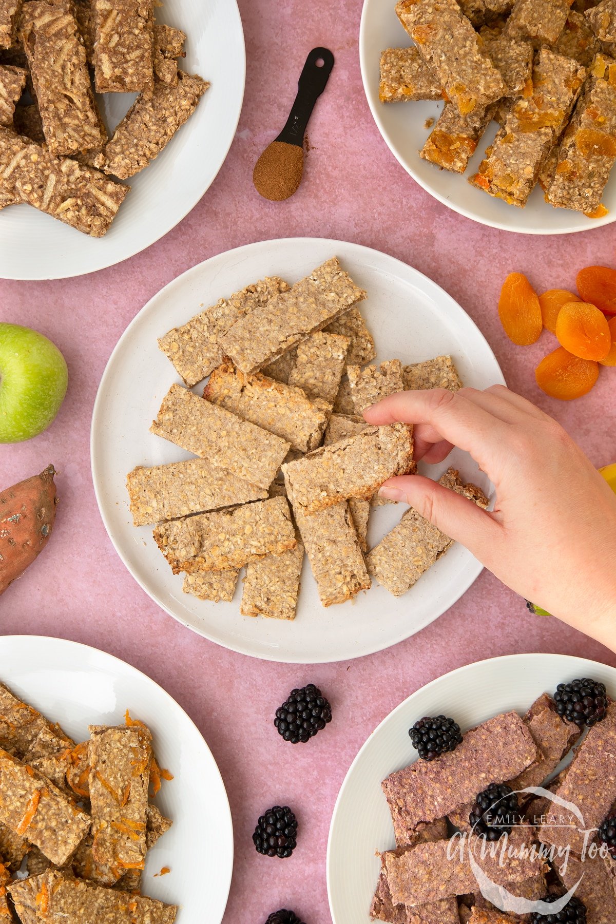 White plate piled with oat fingers. A hand reaches in to take one. Other flavours of oat fingers sit on plates around the edge of the frame.