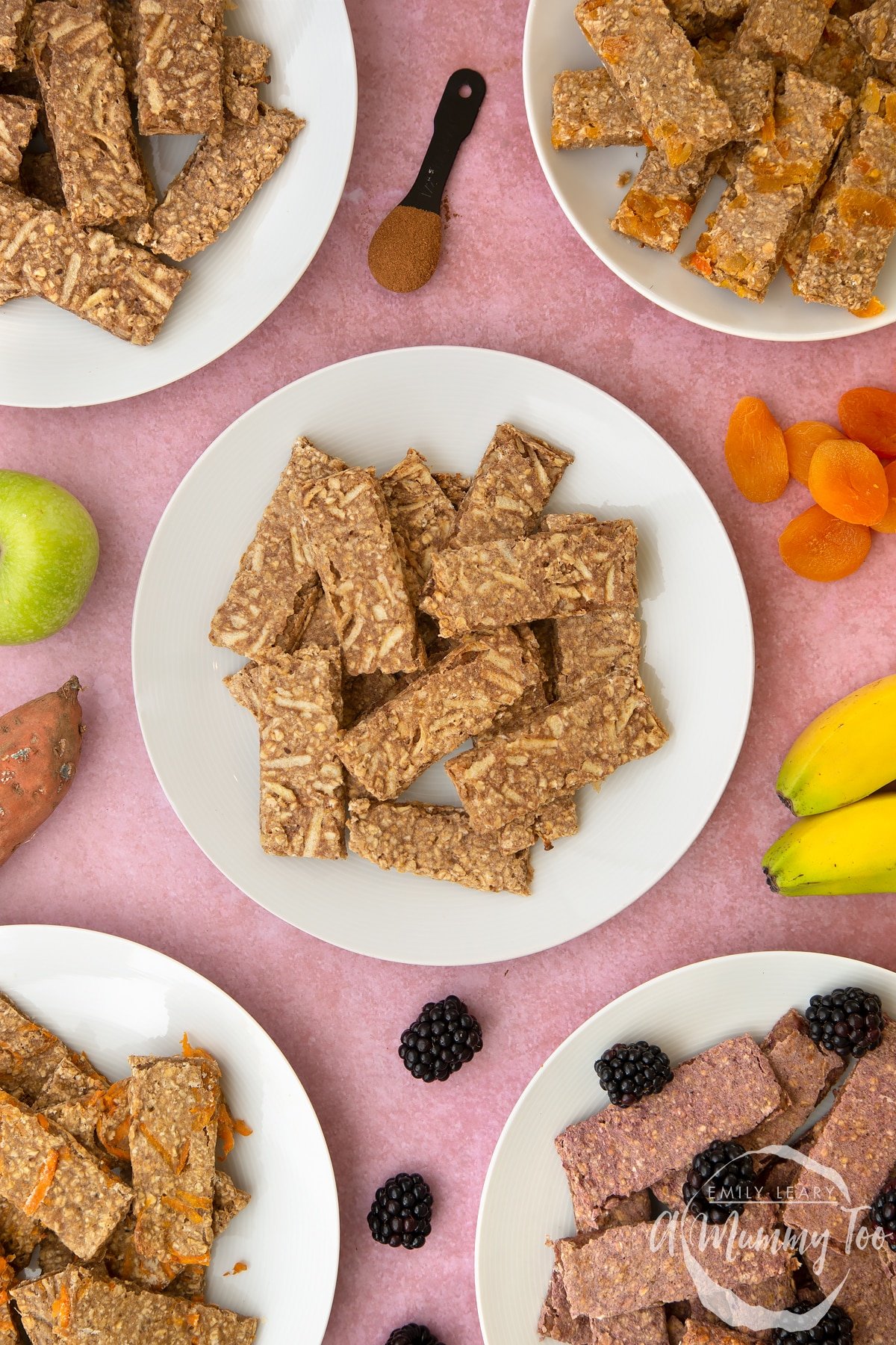 White plate piled with apple and cinnamon oat fingers. Other flavours of oat fingers sit on plates around the edge of the frame.