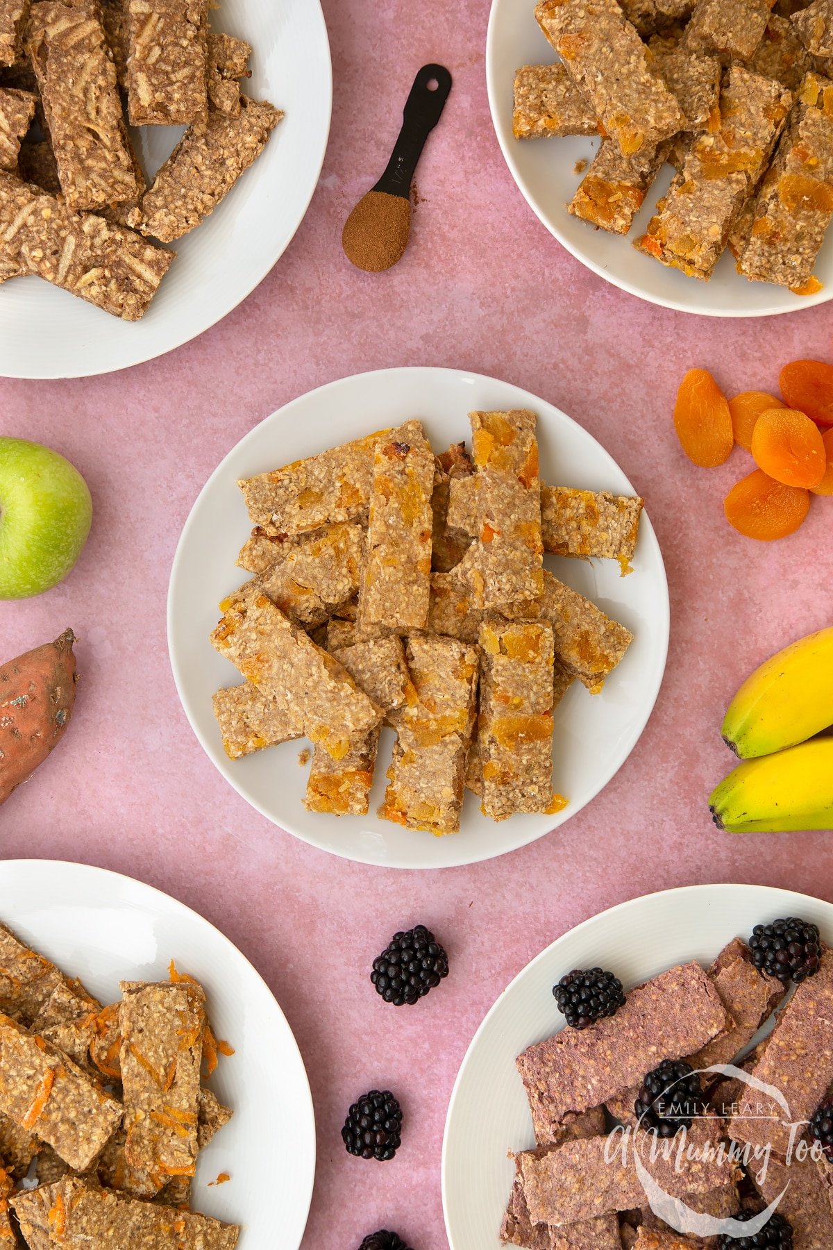 White plate piled with apricot oat fingers. Other flavours of oat fingers sit on plates around the edge of the frame.