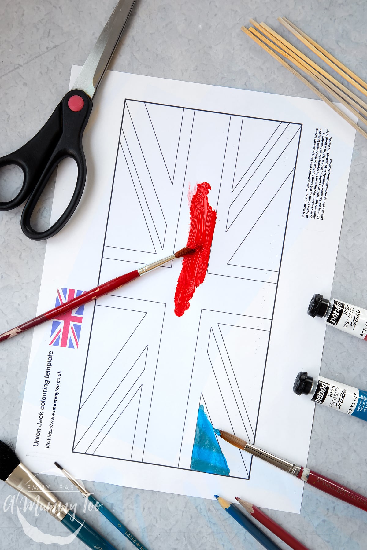 union jack flag printed onto A4 paper with scissors, glue and wooden sticks around the edges and red paint painting the paper