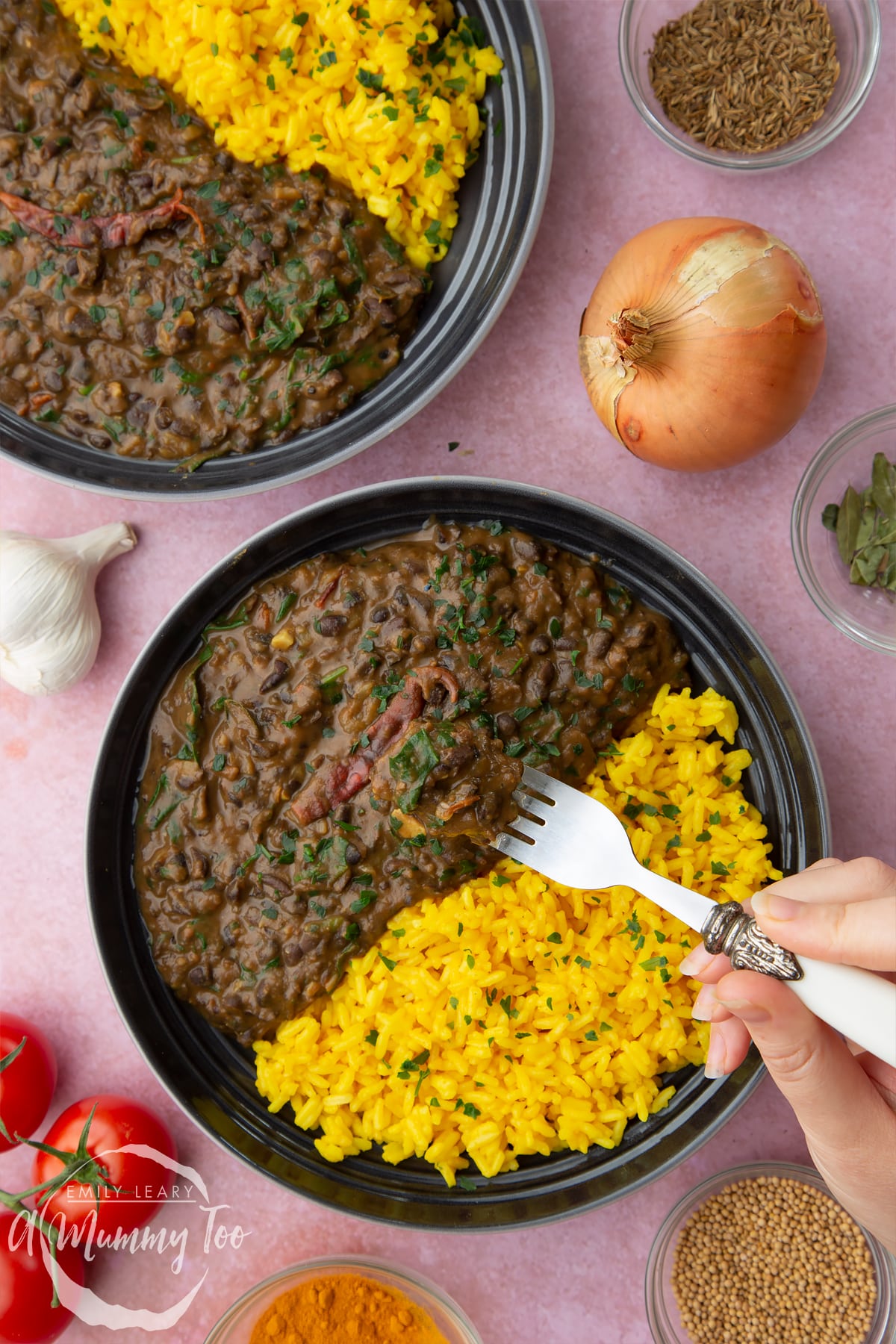 Black bean curry served to two shallow grey bowls with yellow rice. A hand holding a fork reaches in.