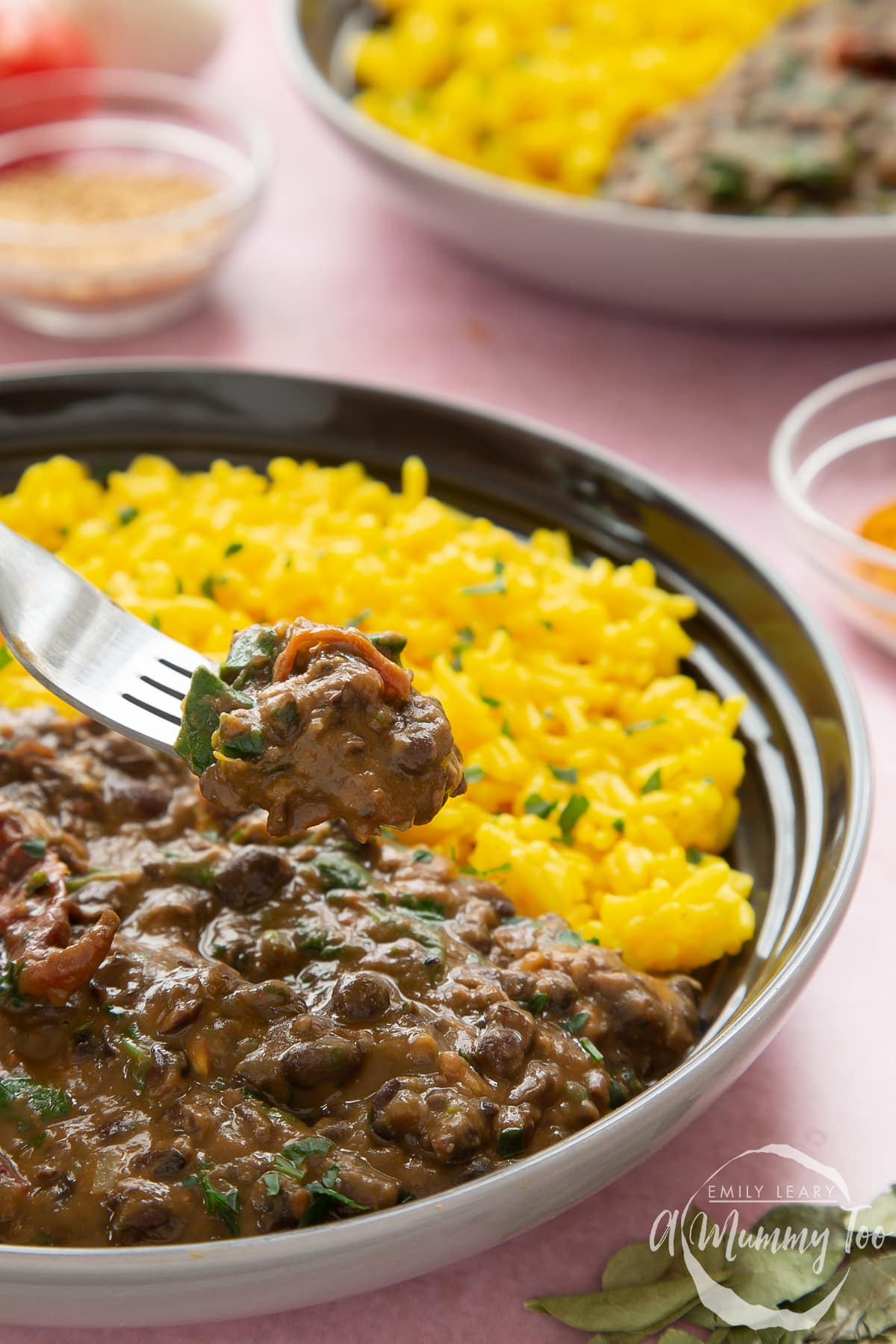 Black bean curry served to a shallow grey bowl with yellow rice scattered with chopped coriander. A fork reaches in.