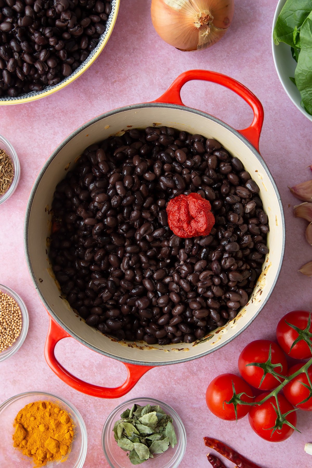 A saucepan containing fried onions, spices and chopped tomatoes with black beans and tomato puree added on top. Ingredients to make black bean curry surround the pan.