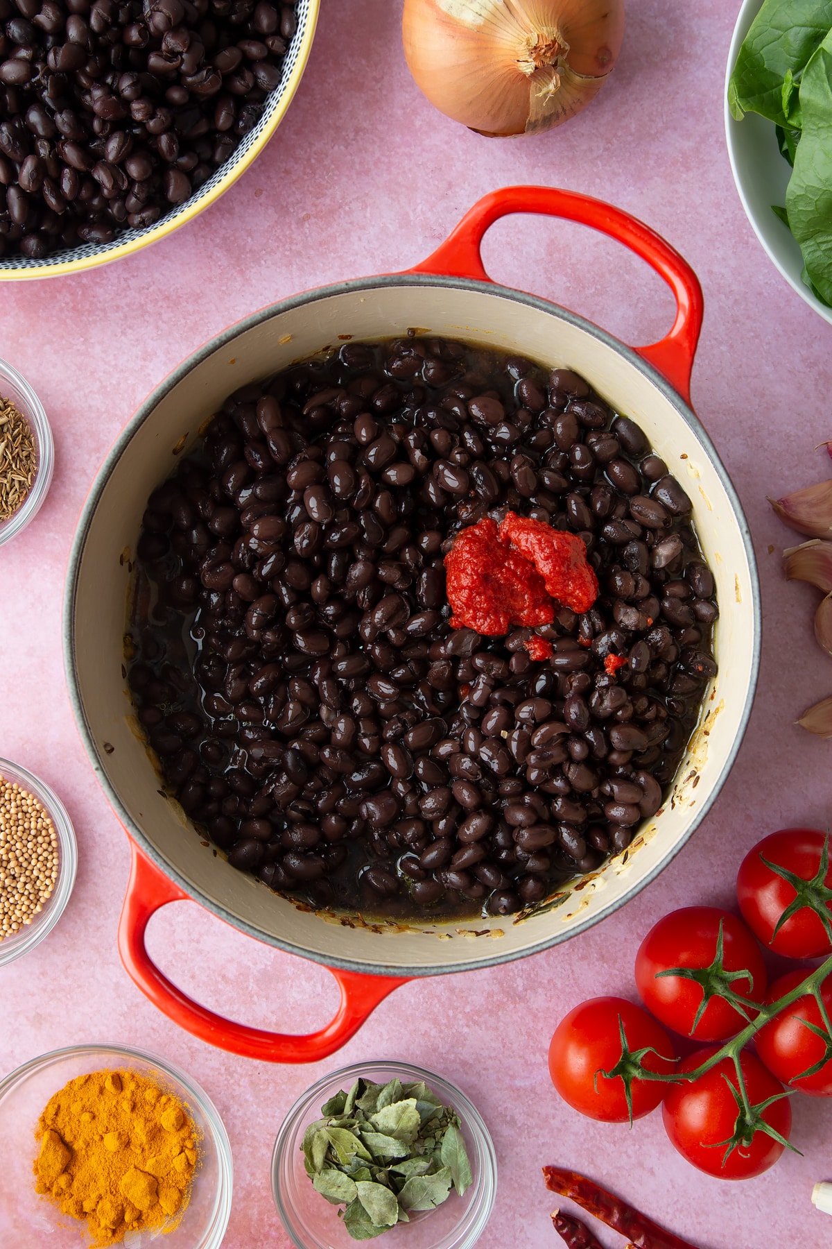 A saucepan containing fried onions, spices and chopped tomatoes with black beans, stock and tomato puree added. Ingredients to make black bean curry surround the pan.