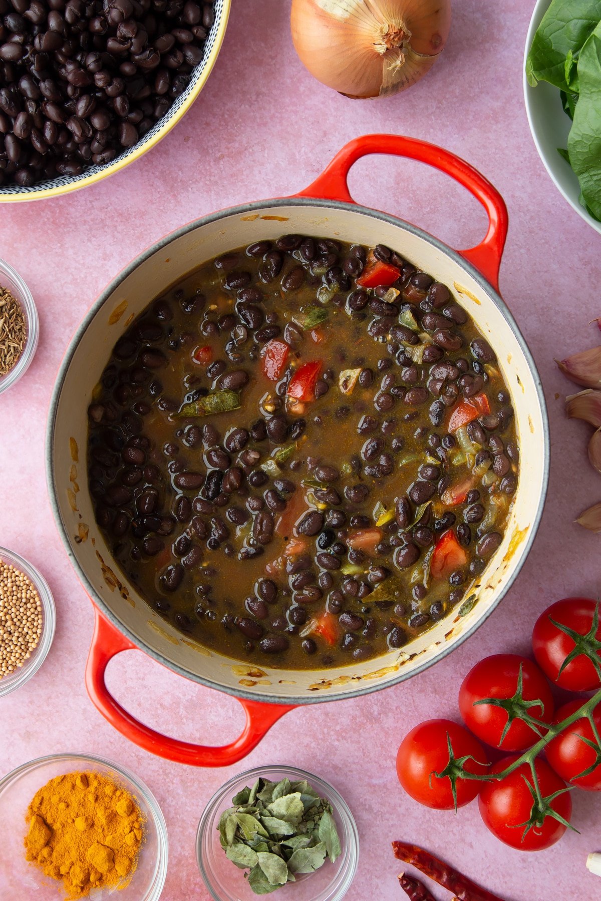 A saucepan containing fried onions, spices, tomatoes, black beans, stock mixed together. Ingredients to make black bean curry surround the pan.