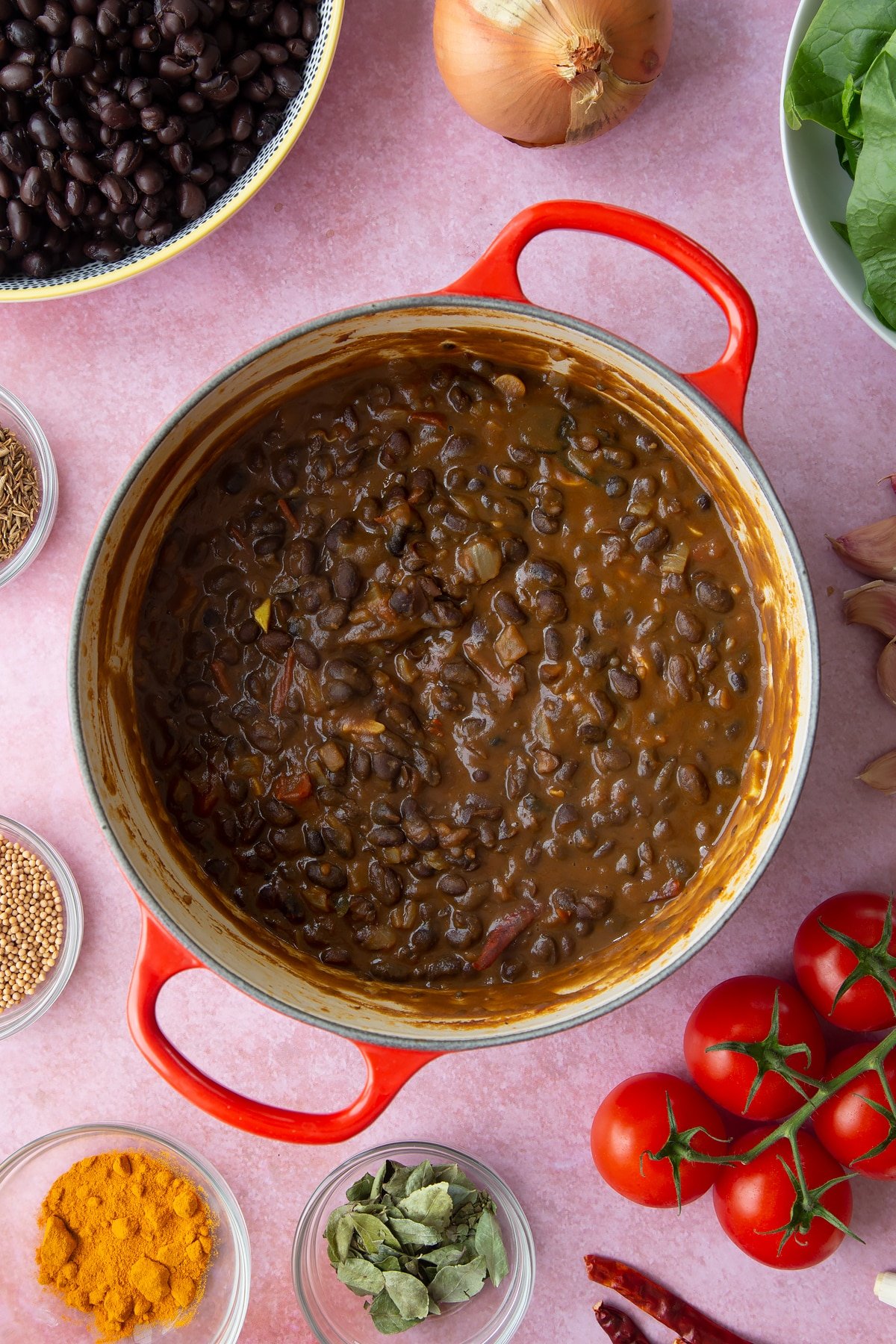 A saucepan containing mashed black beans with fried onions, spices and tomatoes. Ingredients to make black bean curry surround the pan.