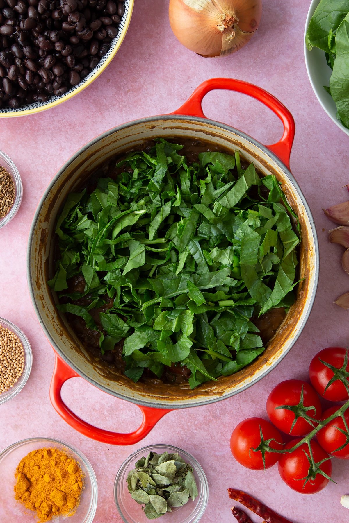 A saucepan containing spiced mashed black beans with shredded spinach on top. Ingredients to make black bean curry surround the pan.