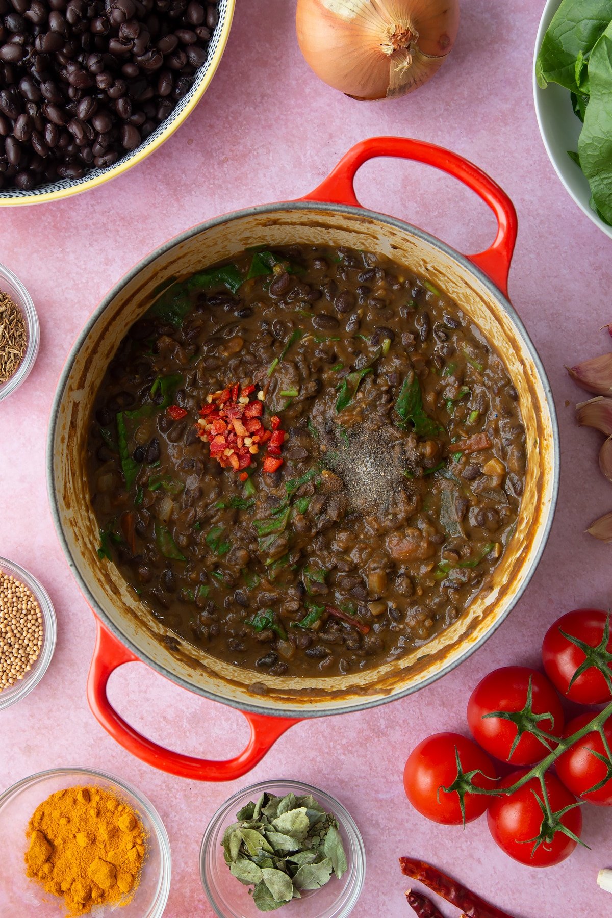 A saucepan containing spiced mashed black beans chopped red chilli on top. Ingredients to make black bean curry surround the pan.