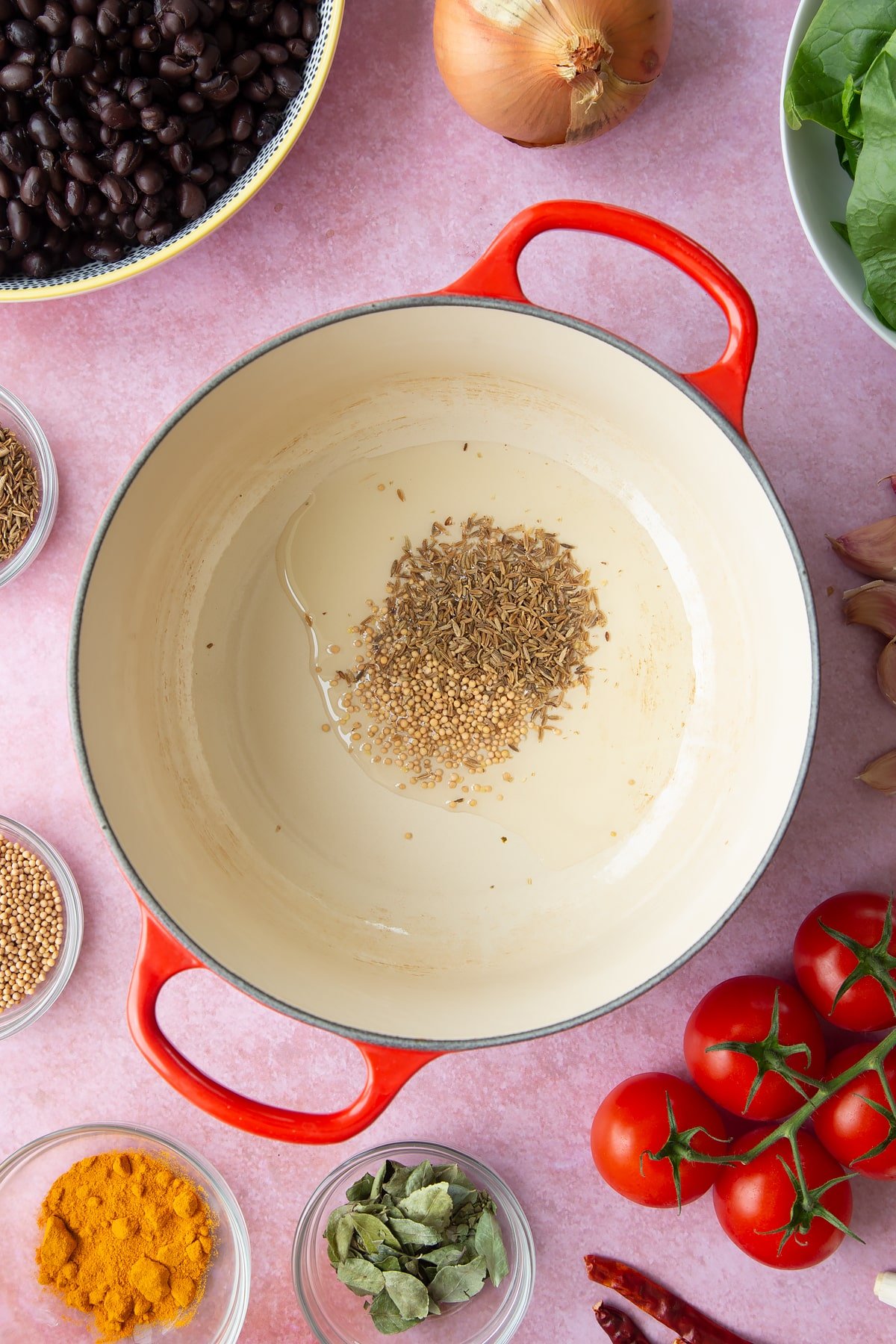 A saucepan containing oil, mustard seeds and cumin seeds. Ingredients to make black bean curry surround the pan.