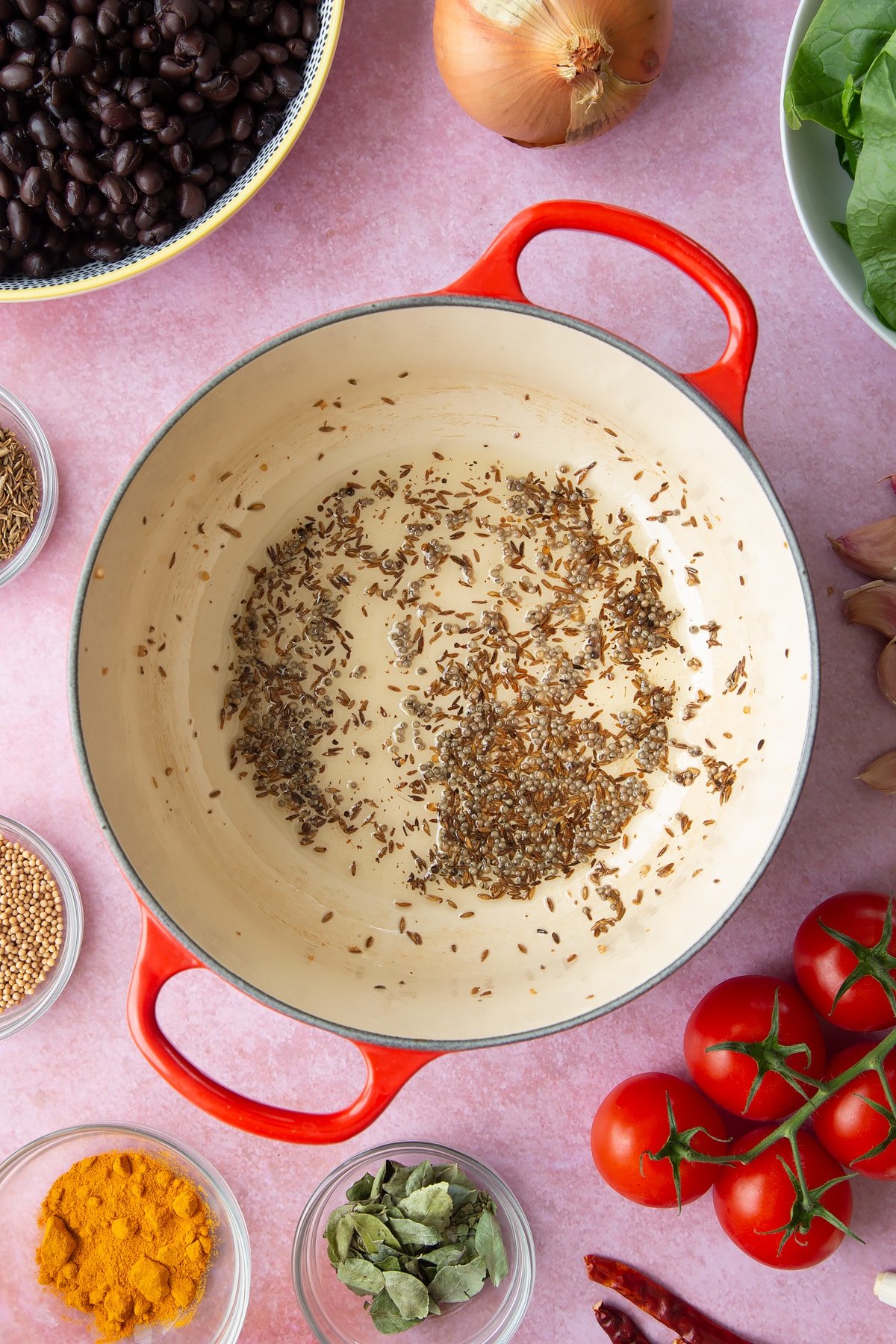 A saucepan containing fried mustard seeds and cumin seeds. Ingredients to make black bean curry surround the pan.