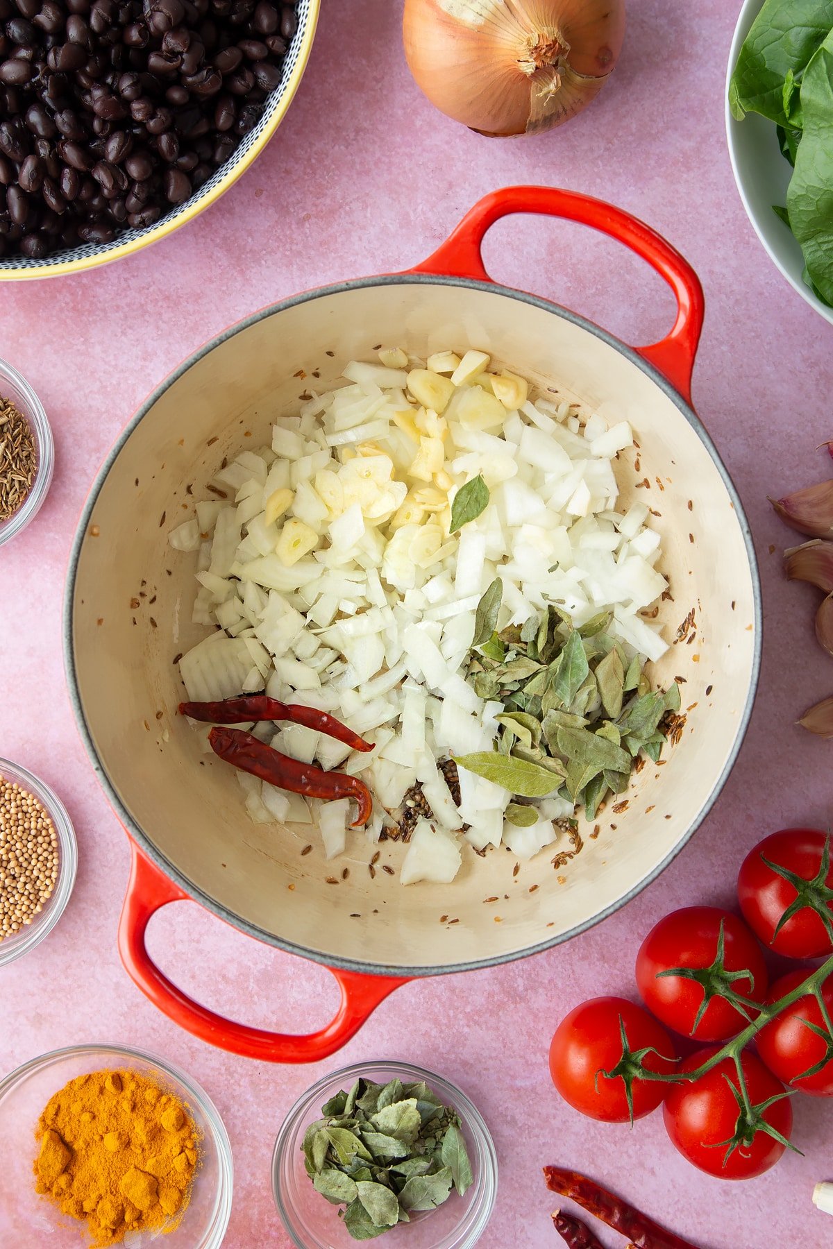 A saucepan containing fried mustard seeds and cumin seeds. Chopped onions and garlic, plus dried curry leaves and red chillies are on top. Ingredients to make black bean curry surround the pan.