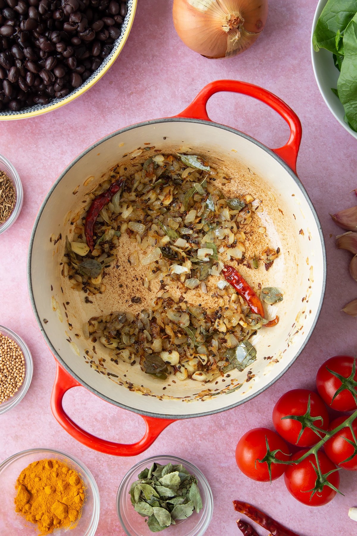 A saucepan containing fried mustard seeds, cumin seeds, onions, garlic, dried curry leaves and red chillies. Ingredients to make black bean curry surround the pan.