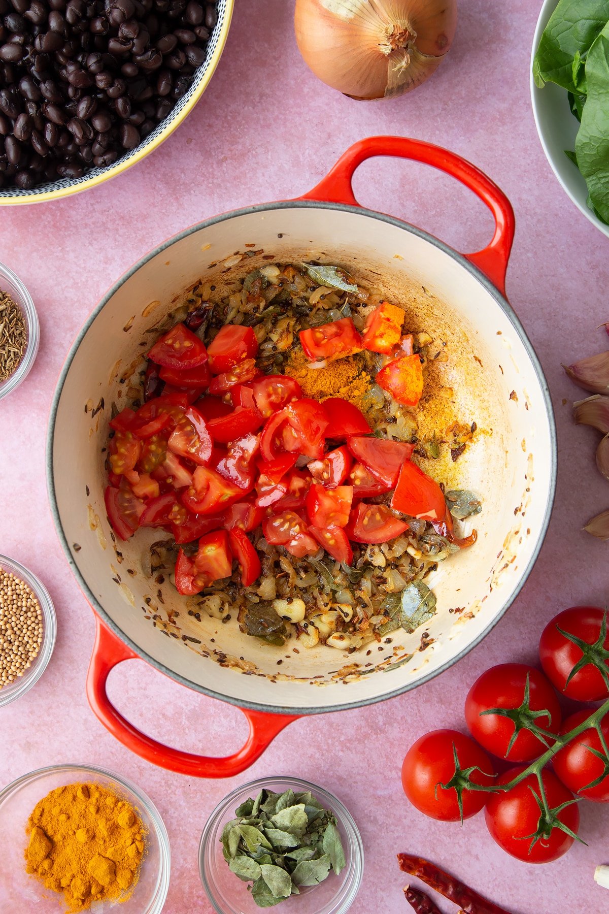 A saucepan containing fried onions and spices with chopped tomatoes and turmeric on top. Ingredients to make black bean curry surround the pan.