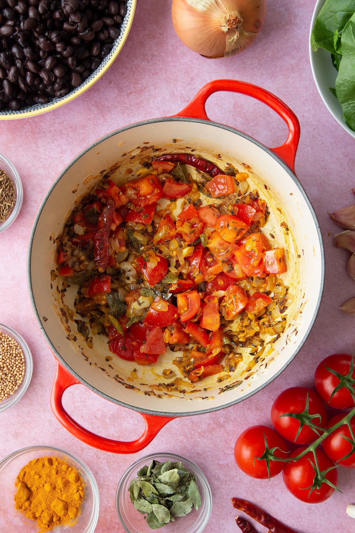 A saucepan containing fried onions, spices and chopped tomatoes. Ingredients to make black bean curry surround the pan.