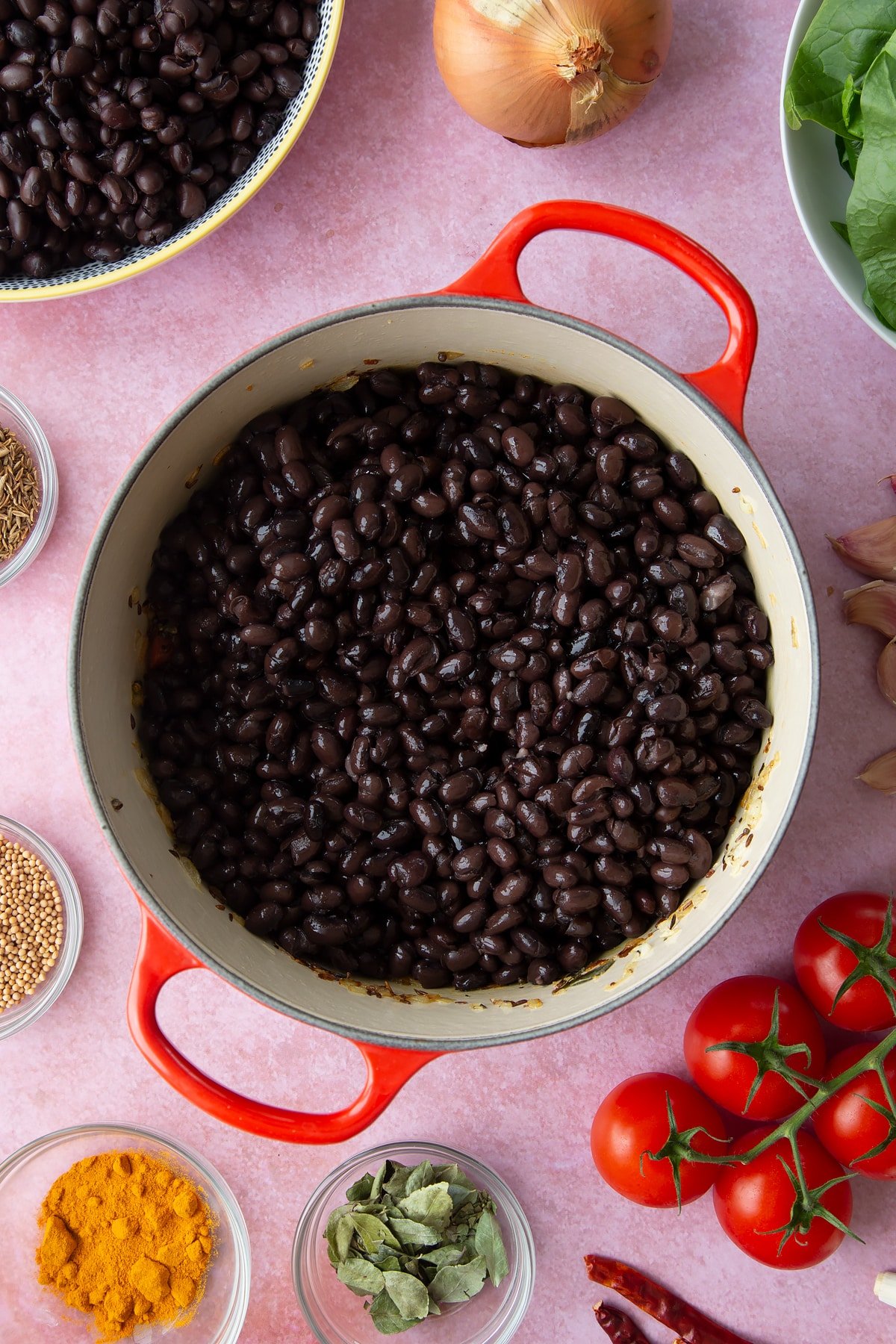 A saucepan containing fried onions, spices and chopped tomatoes with black beans added on top. Ingredients to make black bean curry surround the pan.