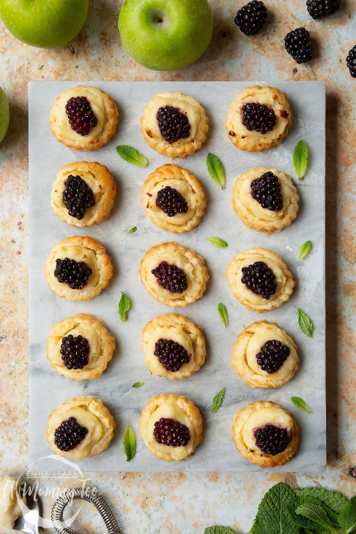 Sixteen blackberry tartlets comprised of a small puff pastry disc topped with sliced apple, pastry cream and a blackberry sit on a marble board with tiny mint leaves scattered around them.