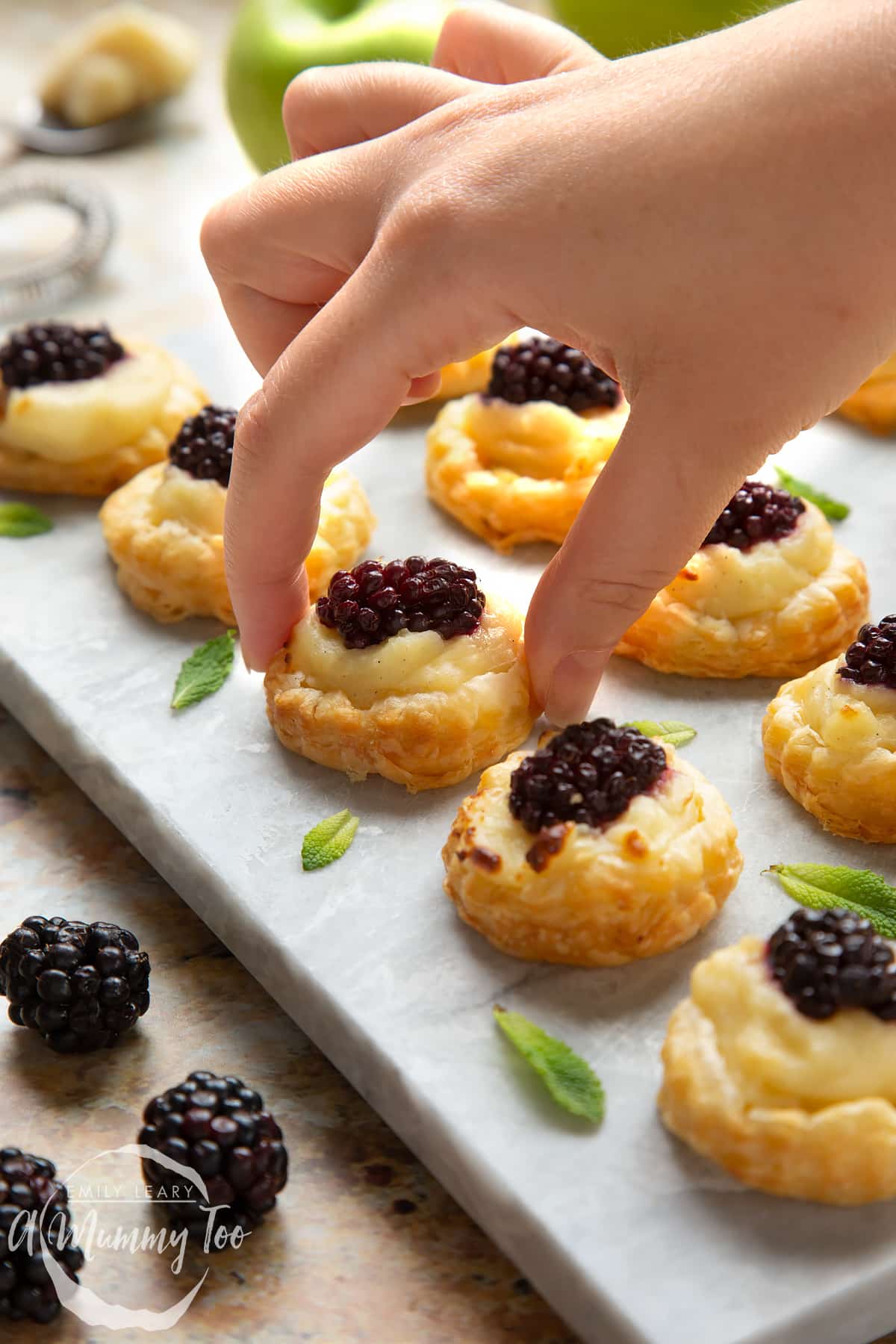 A hand reaches in to take a blackberry tartlet, comprised of a small puff pastry disc topped with sliced apple, pastry cream and a blackberry. More blackberry tartlets sit on the marble board around it.