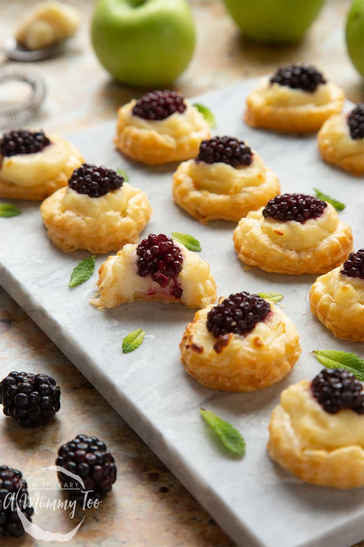 Sixteen blackberry tartlets comprised of a small puff pastry disc topped with sliced apple, pastry cream and a blackberry sit on a marble board with tiny mint leaves scattered around them. One has a bite taken out of it. 