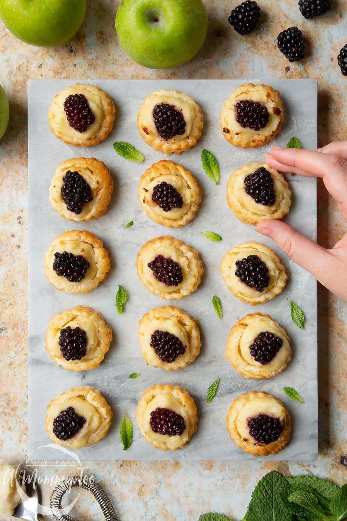 Blackberry tartlets comprised of a small puff pastry disc topped with sliced apple, pastry cream and a blackberry sit on a marble board with tiny mint leaves scattered around them. A hand holds one of the tarts. 