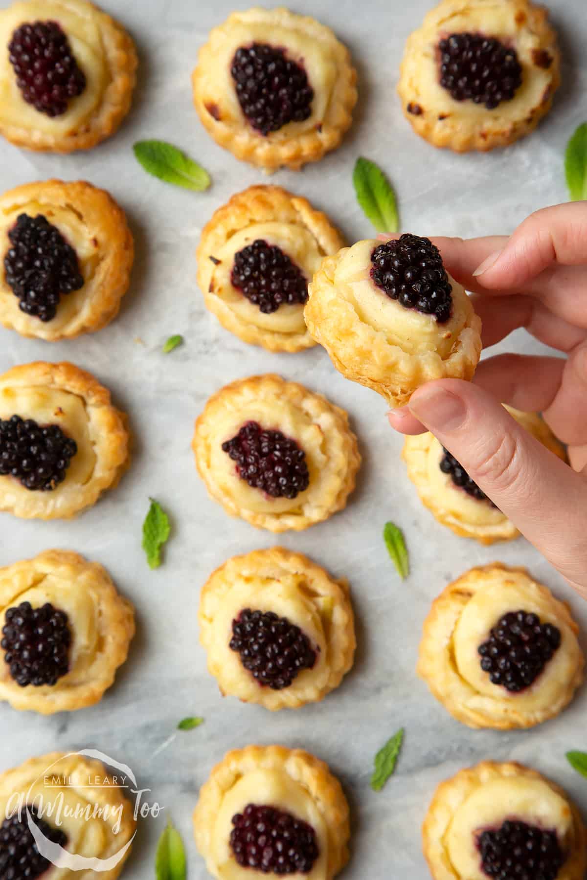 A hand holds a blackberry tartlet, comprised of a small puff pastry disc topped with sliced apple, pastry cream and a blackberry. More blackberry tartlets sit on the marble board in the background.