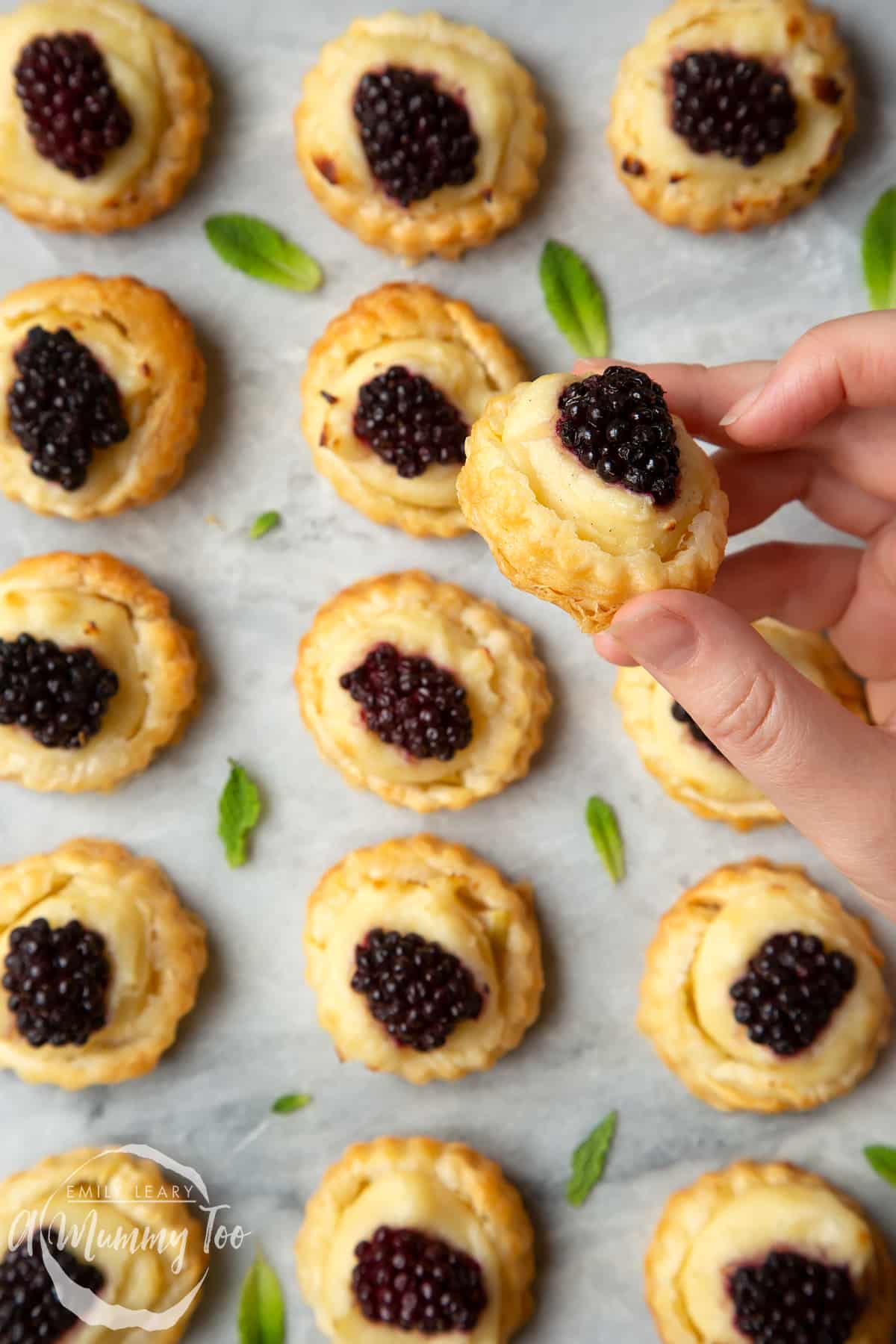 Sixteen blackberry tartlets comprised of a small puff pastry disc topped with sliced apple, pastry cream and a blackberry sit on a marble board with tiny mint leaves scattered around them. A hand holds one of the tarts. 