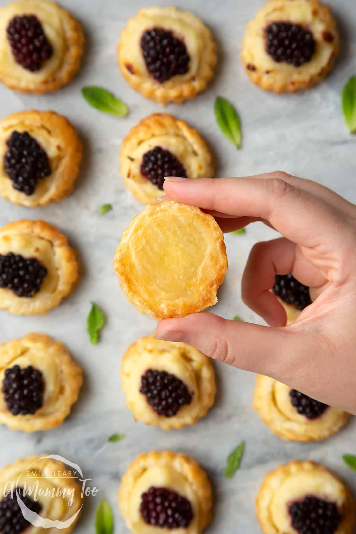 A hand holding a blackberry tartlet, showing the golden underside. More blackberry tartlets comprised of a small puff pastry disc topped with sliced apple, pastry cream and a blackberry sit on the marble board with tiny mint leaves scattered around them.