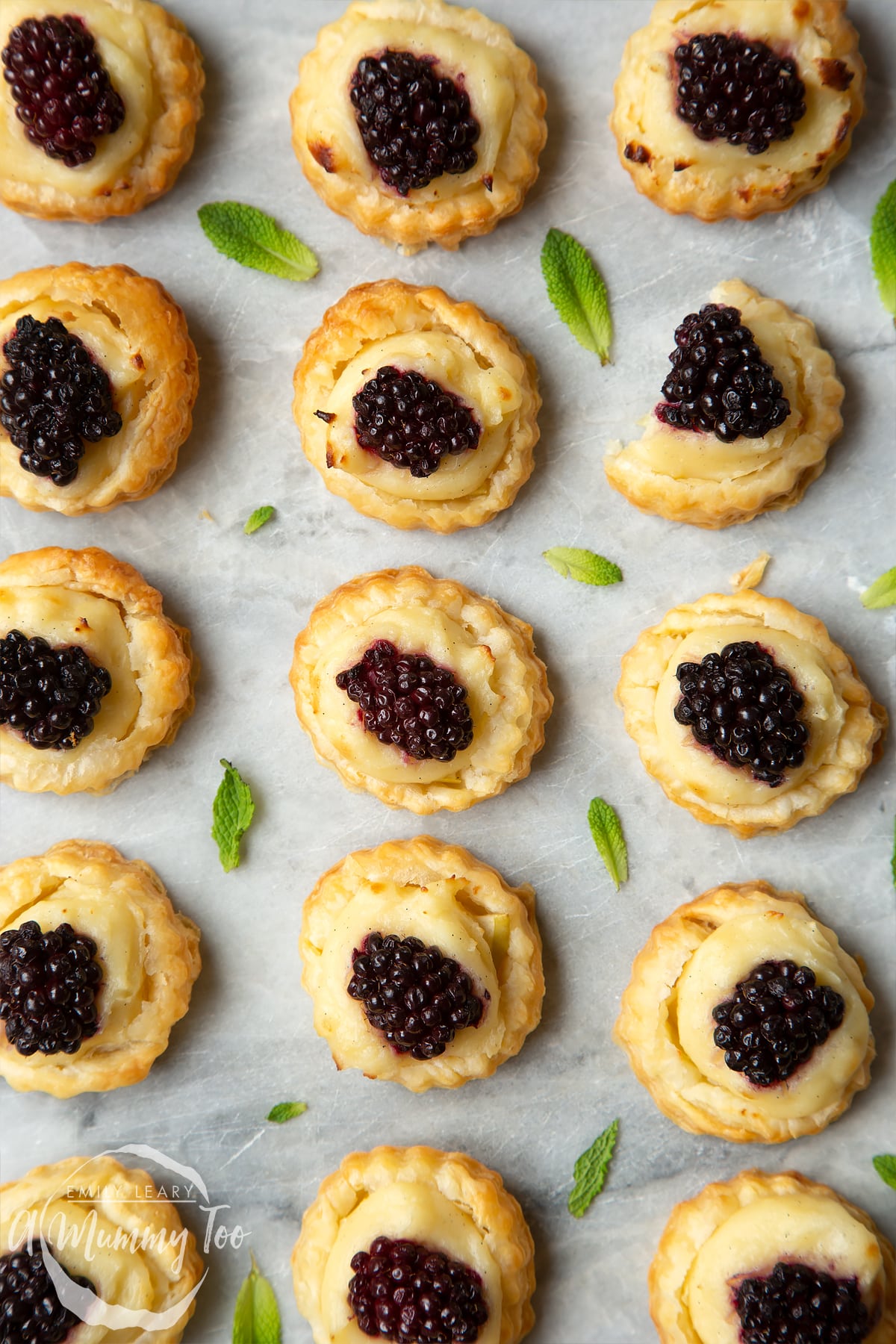 16 blackberry tartlets comprised of a small puff pastry disc topped with sliced apple, pastry cream and a blackberry sit on the marble board with tiny mint leaves scattered around them.