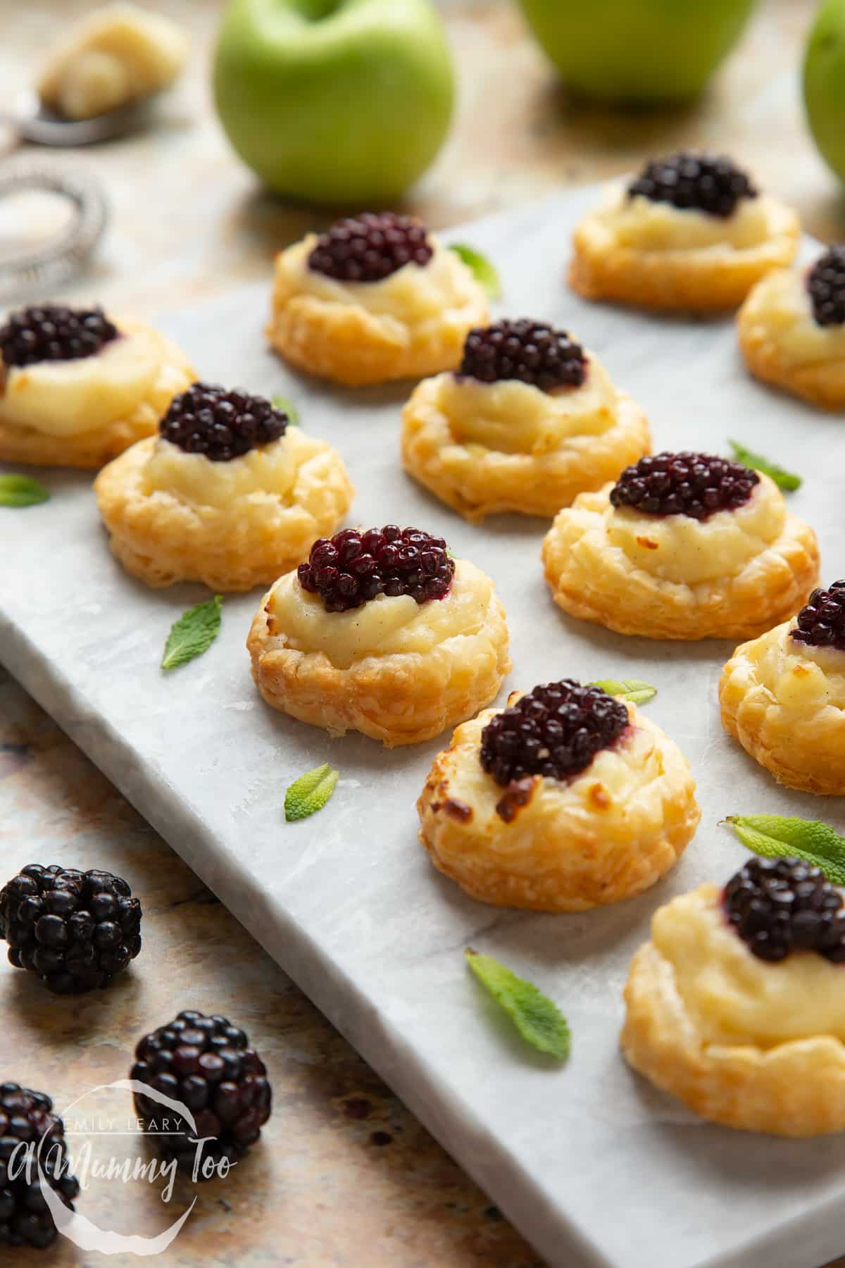 Blackberry tartlets comprised of a small puff pastry disc topped with sliced apple, pastry cream and a blackberry sit on a marble board with tiny mint leaves scattered around them. 