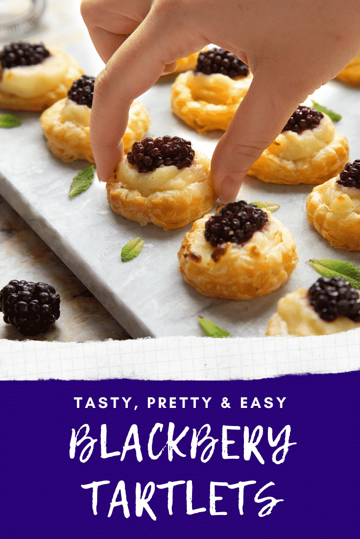 Blackberry tartlets on a marble board with tiny mint leaves scattered around them. A hand holds one of the tarts. Caption reads: tasty, pretty, easy blackberry tartlets.