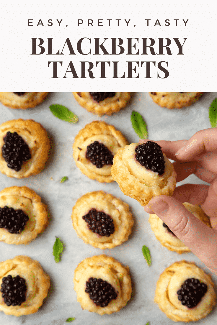 Blackberry tartlets on a marble board with tiny mint leaves scattered around them. A hand holds one of the tarts. Caption reads: easy, pretty, tasty blackberry tartlets.