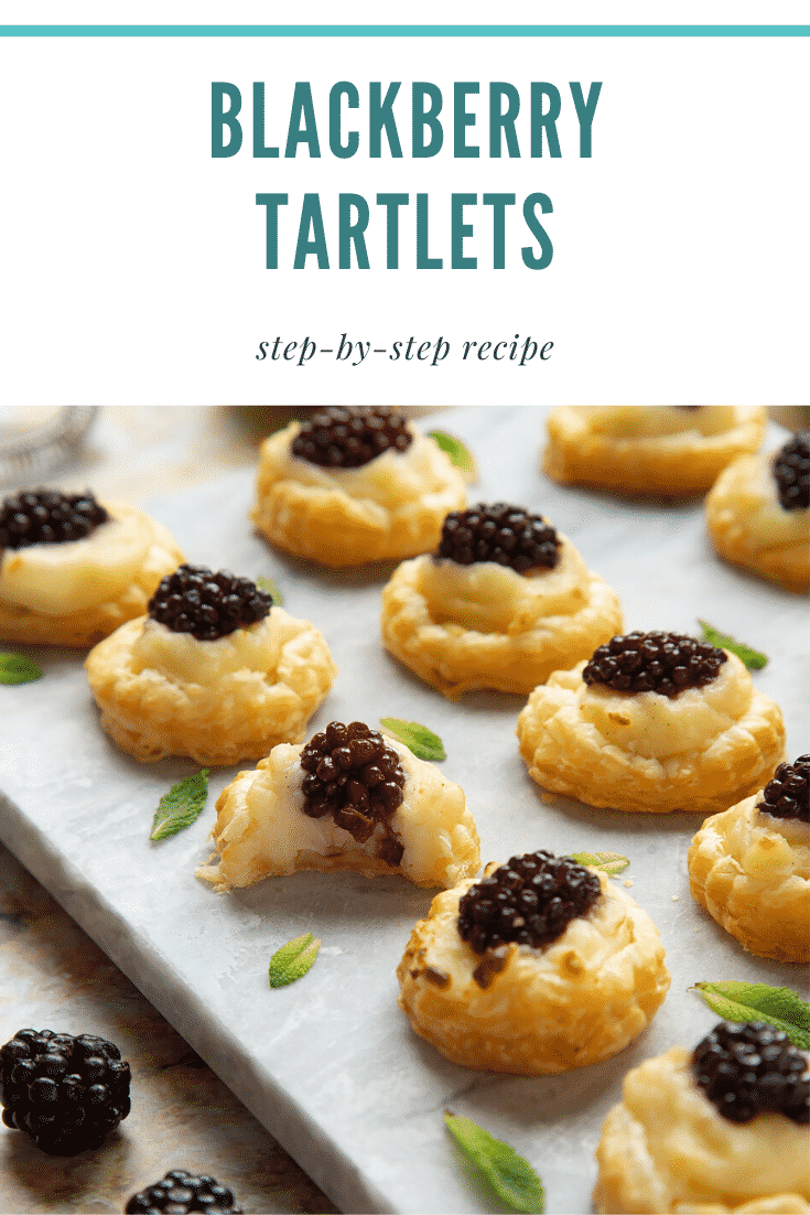 Blackberry tartlets on a marble board with tiny mint leaves scattered around them. A bite is taken out of one. Caption reads: blackberry tartlets step-by-step recipe.