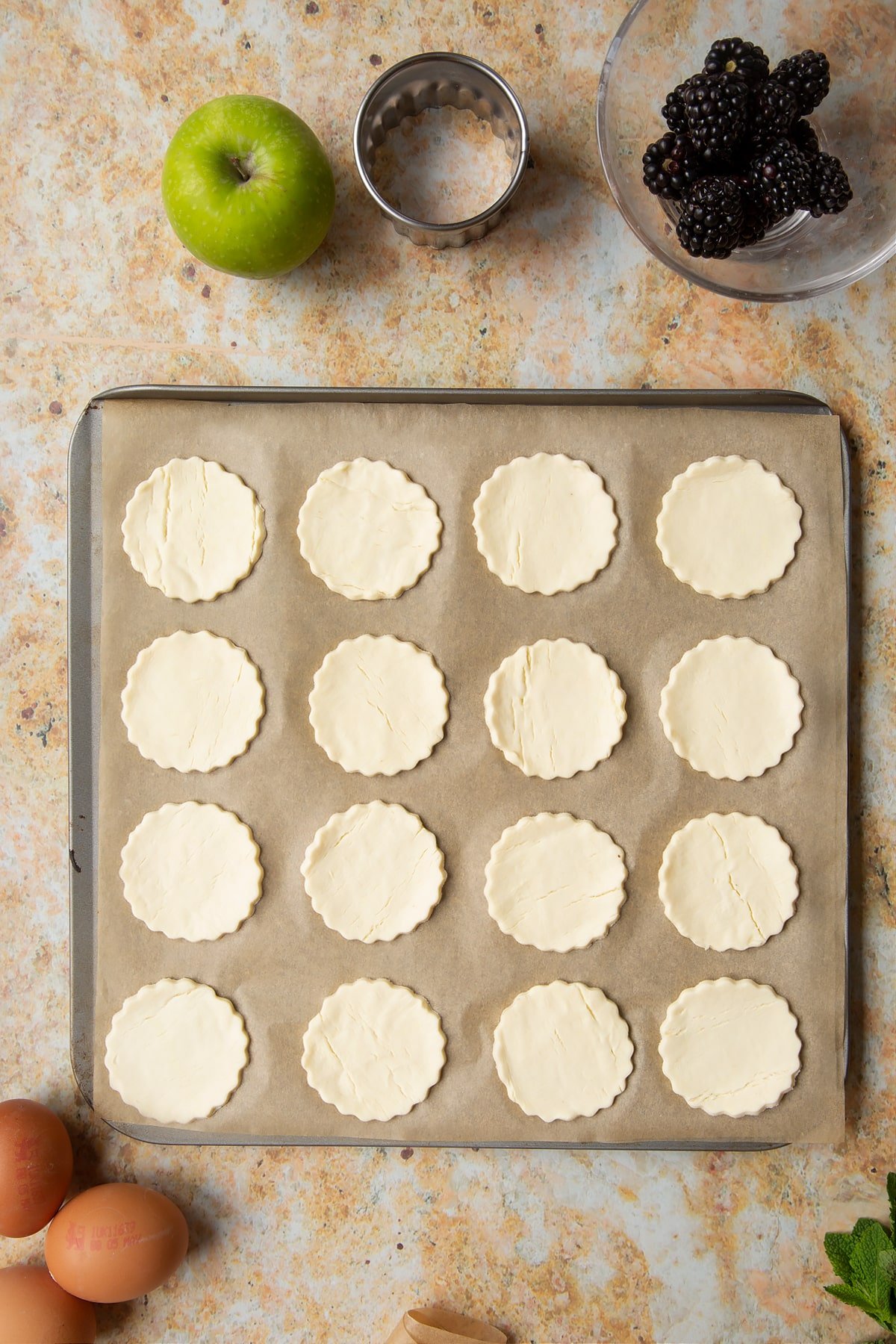 16 raw puff pastry discs sit on a lined baking sheet, ready to be baked.