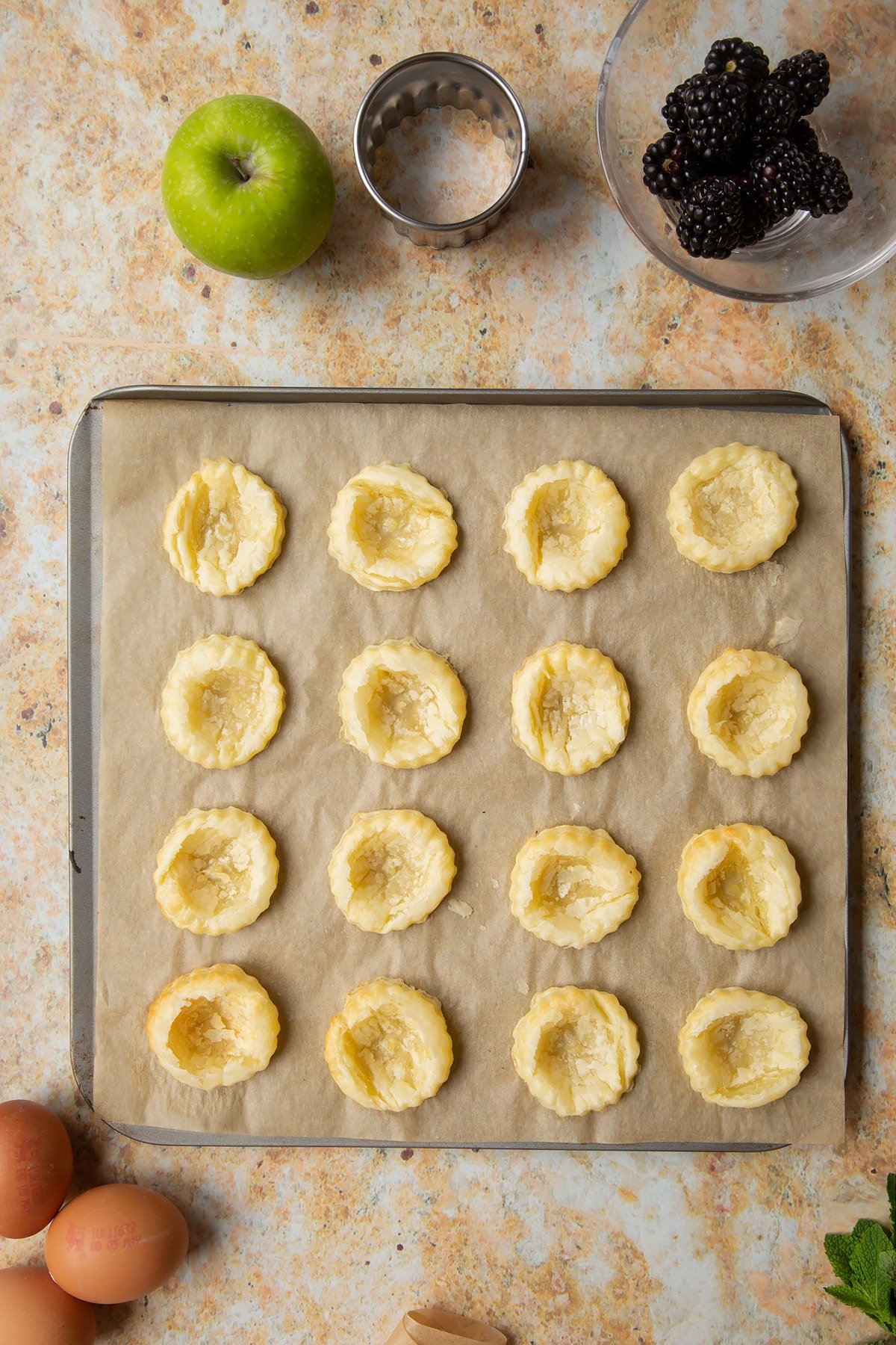 16 puff pastry discs with the middles pressed in sit on a lined baking sheet.