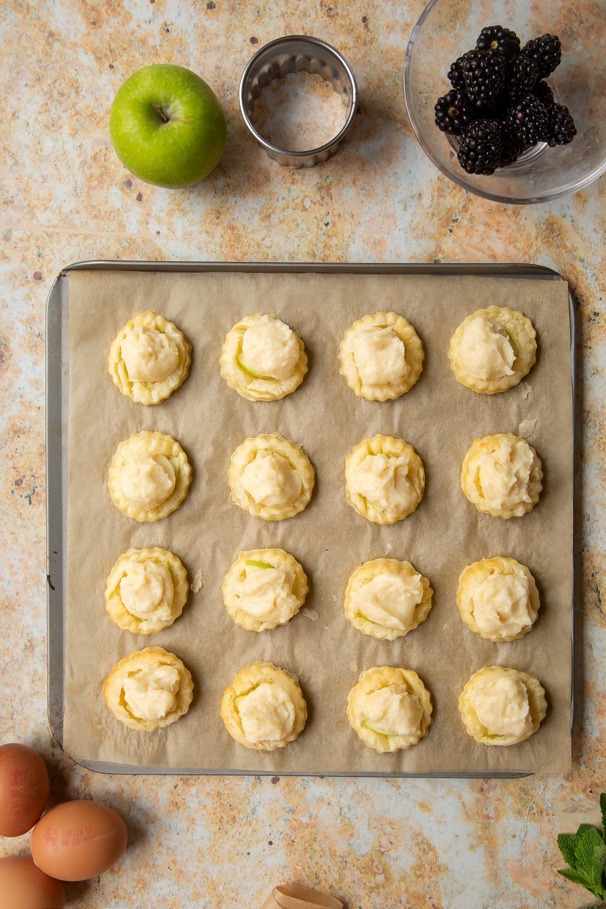 16 puff pastry discs topped with sliced apple and pastry cream sit on a lined baking sheet.