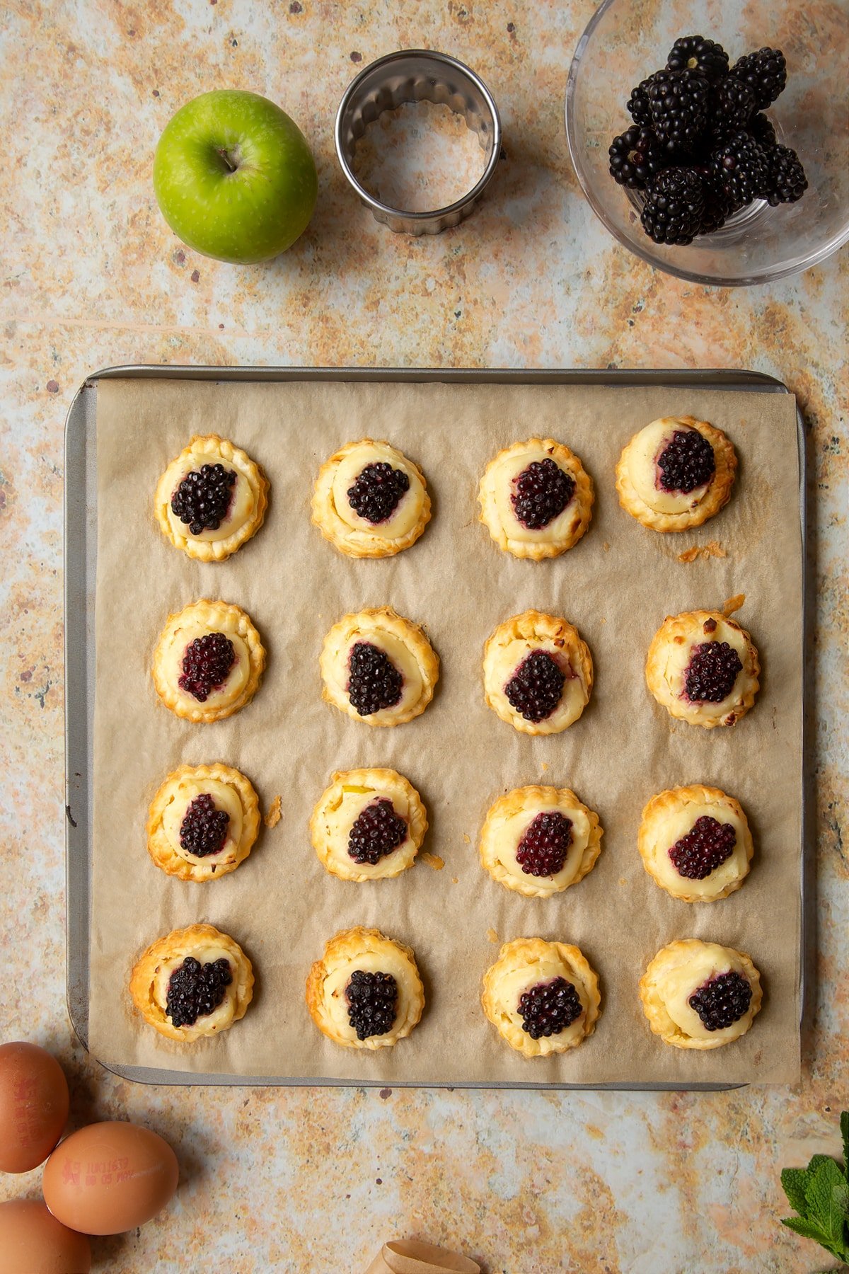 16 freshly baked blackberry tartlets comprised of a small puff pastry disc topped with sliced apple, pastry cream and a blackberry sit on a lined baking sheet.