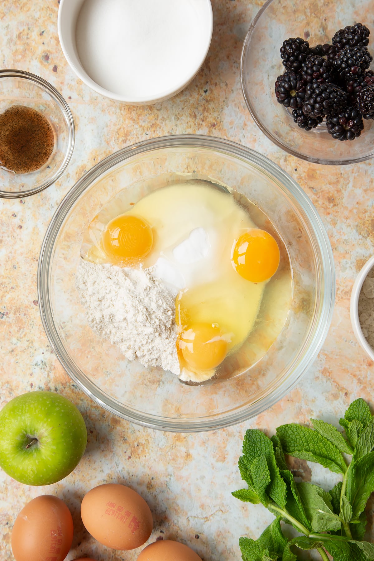 A bowl filled with eggs, flour, sugar and vanilla, surrounded by ingredients to make blackberry tartlets.