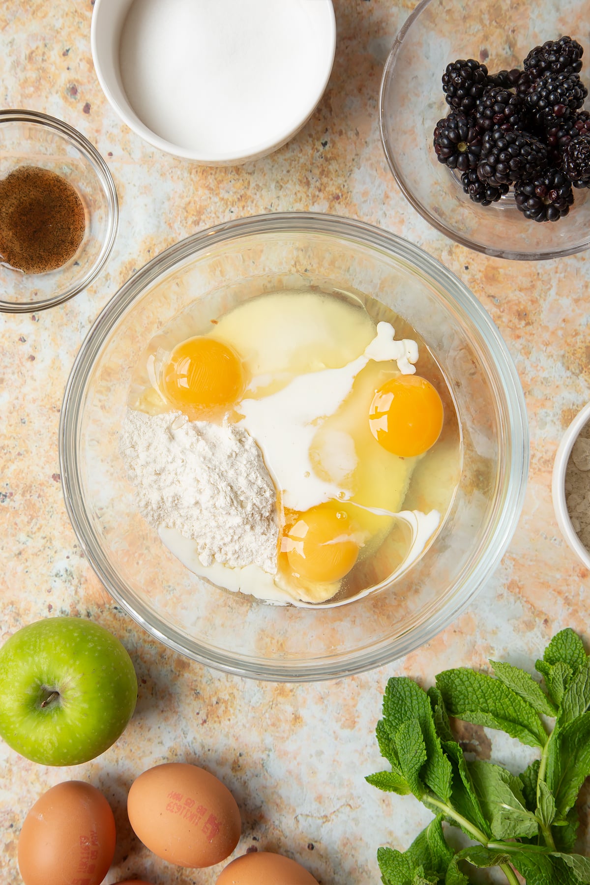 A bowl filled with eggs, flour, sugar, vanilla and a dash of milk, surrounded by ingredients to make blackberry tartlets.