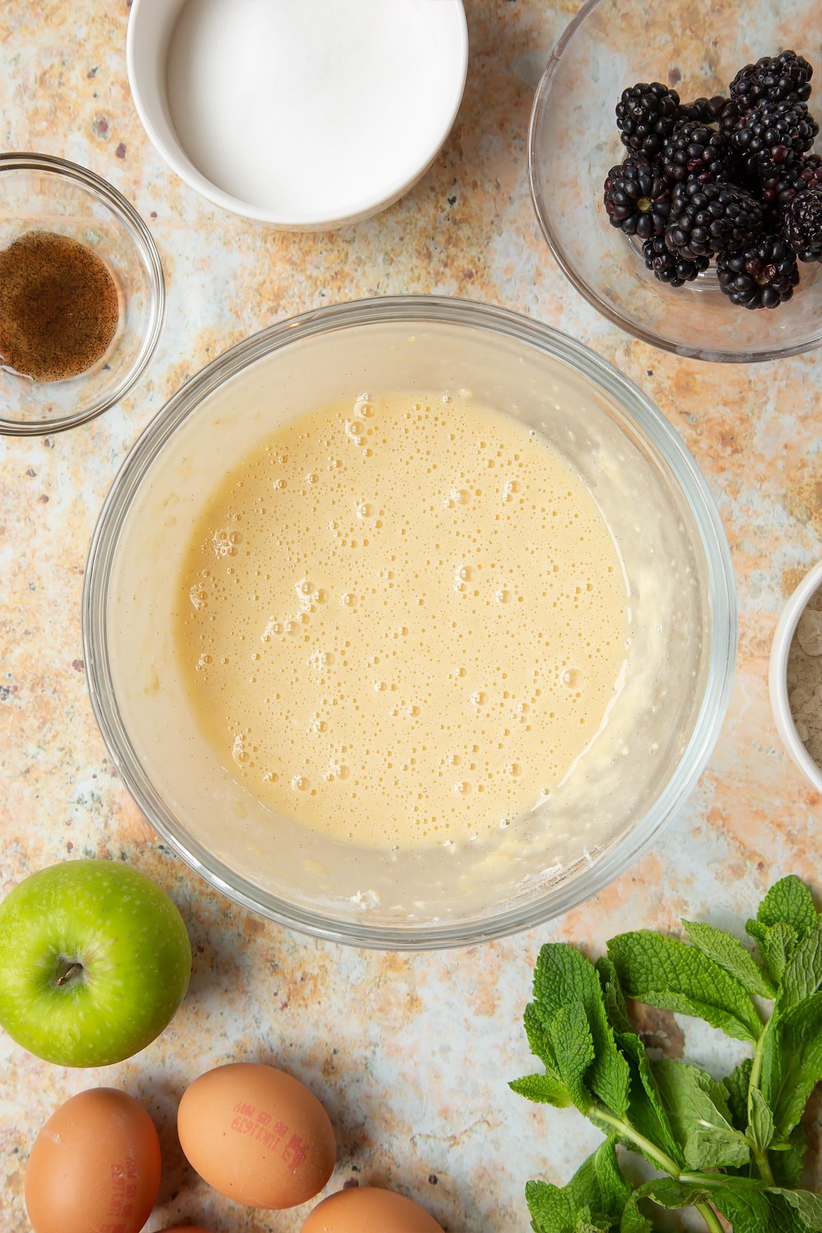 A bowl filled with eggs, flour, sugar, vanilla and a dash of milk whisked together, surrounded by ingredients to make blackberry tartlets.