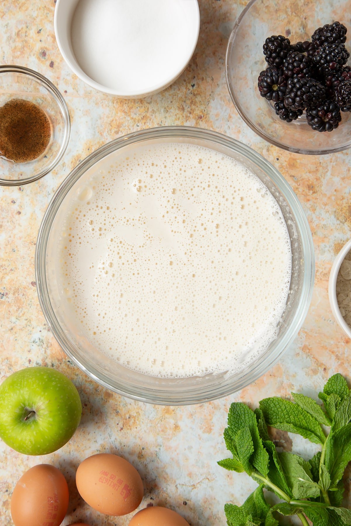 A bowl filled with milk, eggs, flour, sugar and vanilla, surrounded by ingredients to make blackberry tartlets.