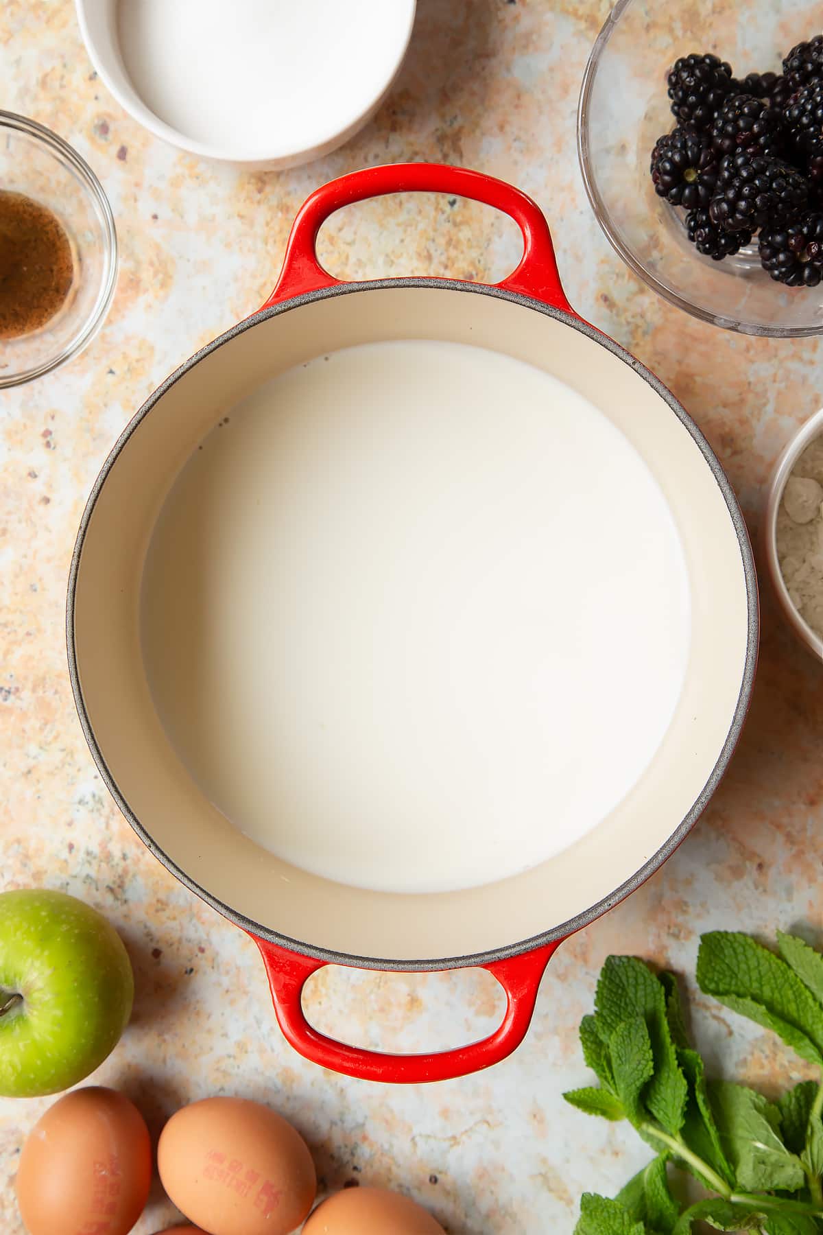 A saucepan filled with milk, surrounded by ingredients to make blackberry tartlets.