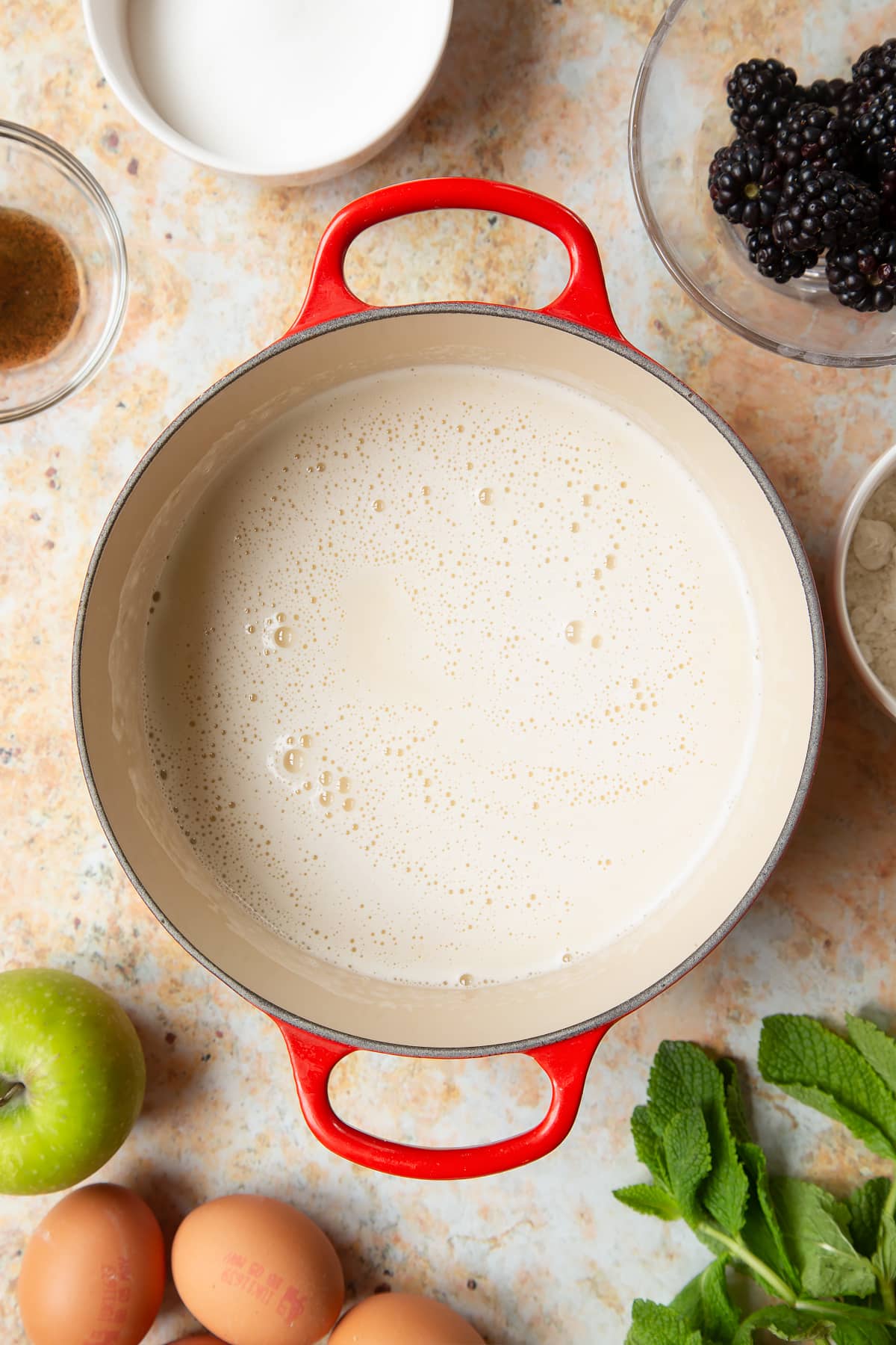 A saucepan filled with milk, eggs, flour, sugar and vanilla, surrounded by ingredients to make blackberry tartlets.