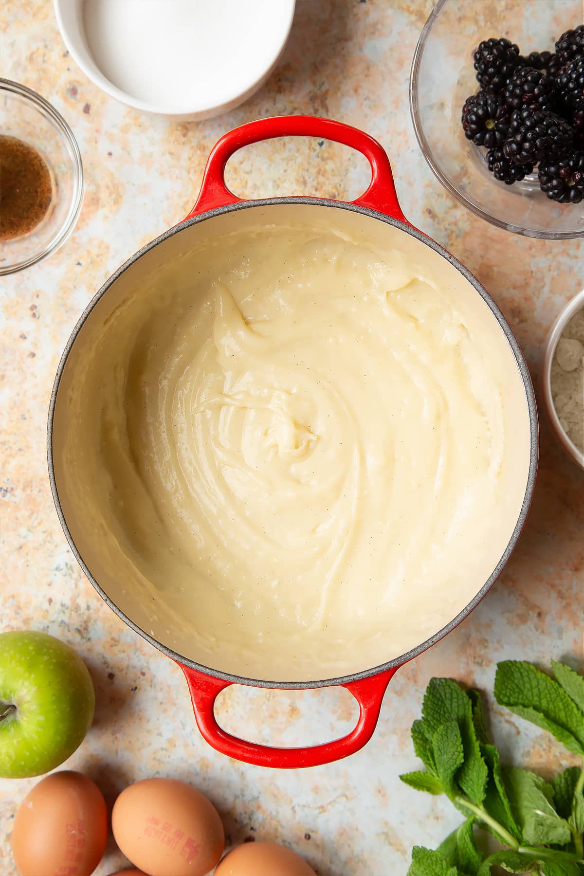 A saucepan filled with pastry cream, surrounded by ingredients to make blackberry tartlets.