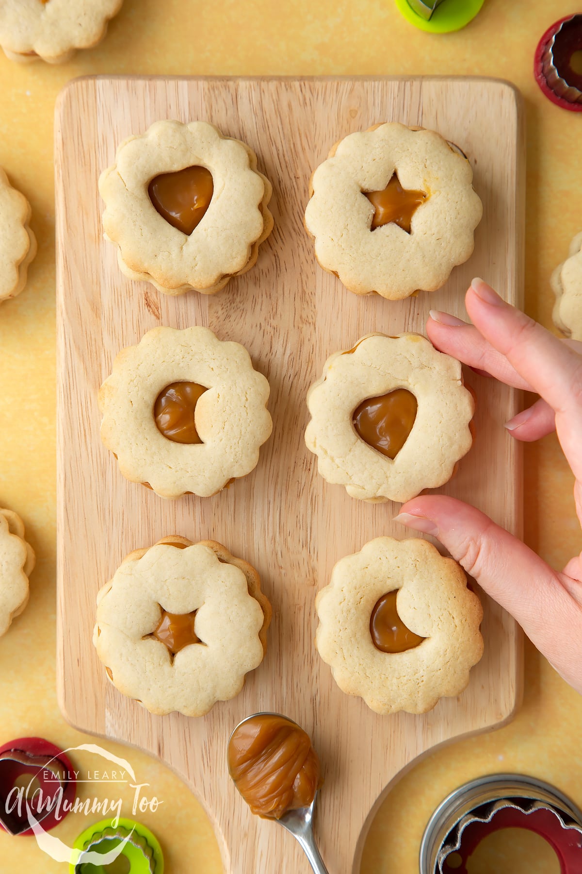 Overhead shot of a hand touching a caramel sandwich cookie served on a wooden plate with a mummy too logo in the lower-left corner