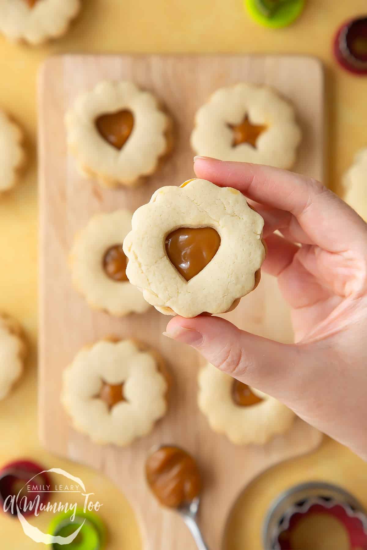 Hand holding a caramel sandwich cookies with additional caramel sandwich cookies in the background. 