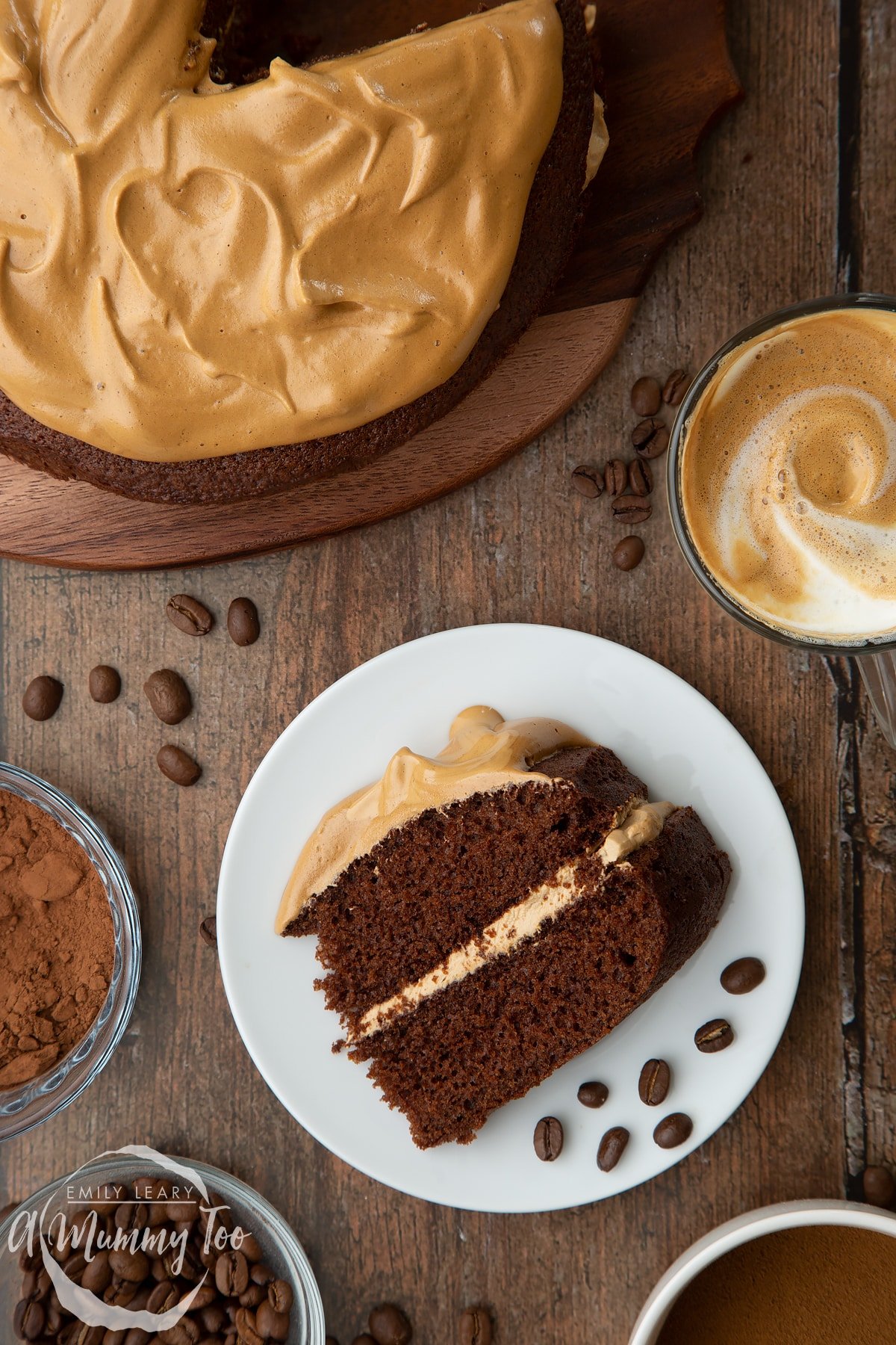 A slice of dalgona coffee cake on a white plate with a few coffee beans. A cup of dalgona coffee, cocoa and coffee beans are shown to the sides.