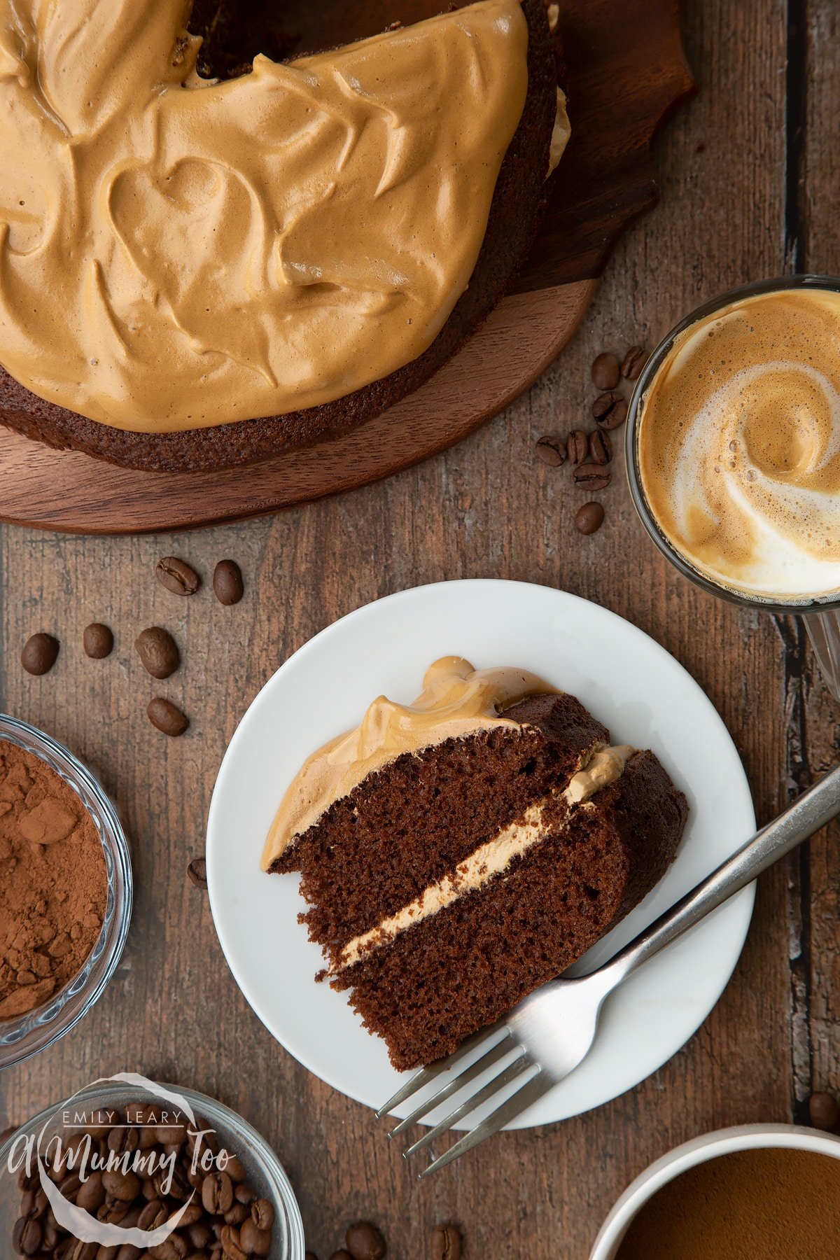 A slice of dalgona coffee cake on a white plate with a fork. A cup of dalgona coffee, cocoa and coffee beans are shown to the sides.