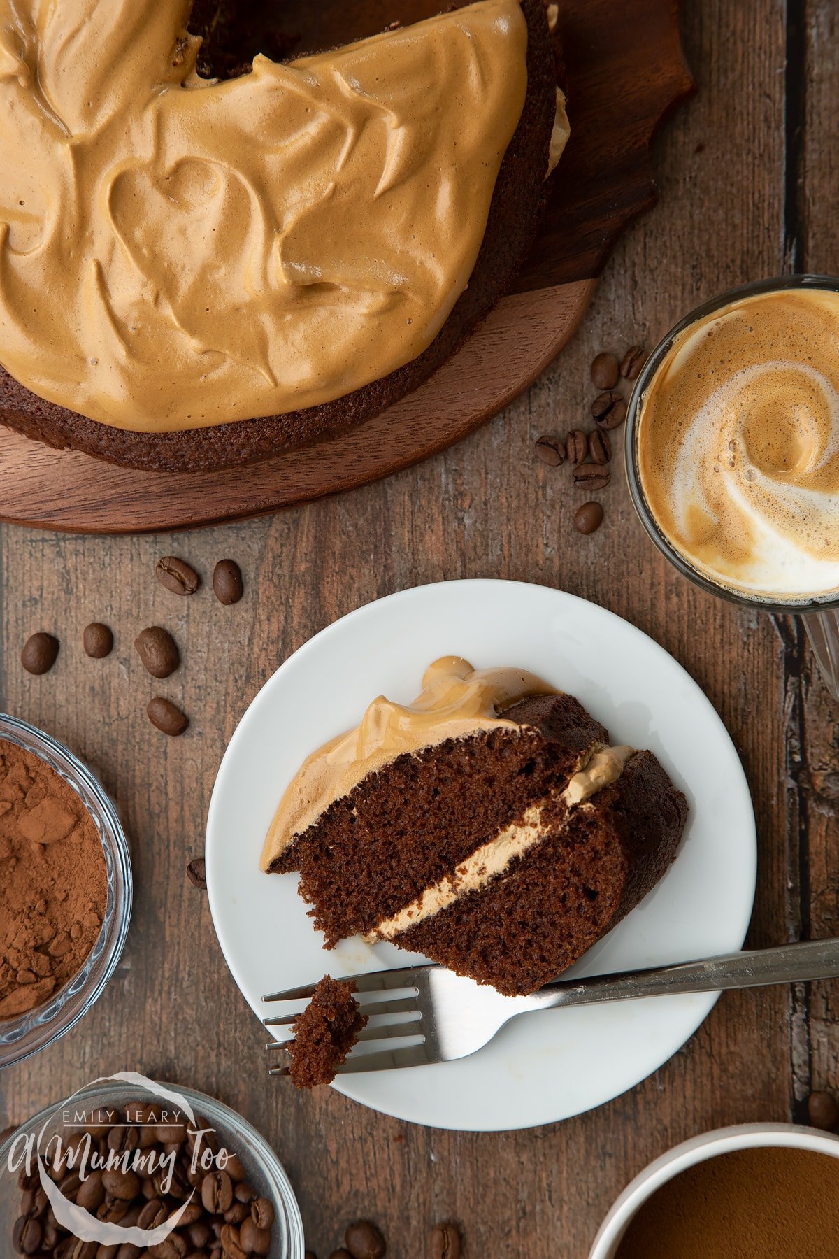 A slice of dalgona coffee cake on a white plate. A fork has taken a piece. A cup of dalgona coffee, cocoa and coffee beans are shown to the sides.