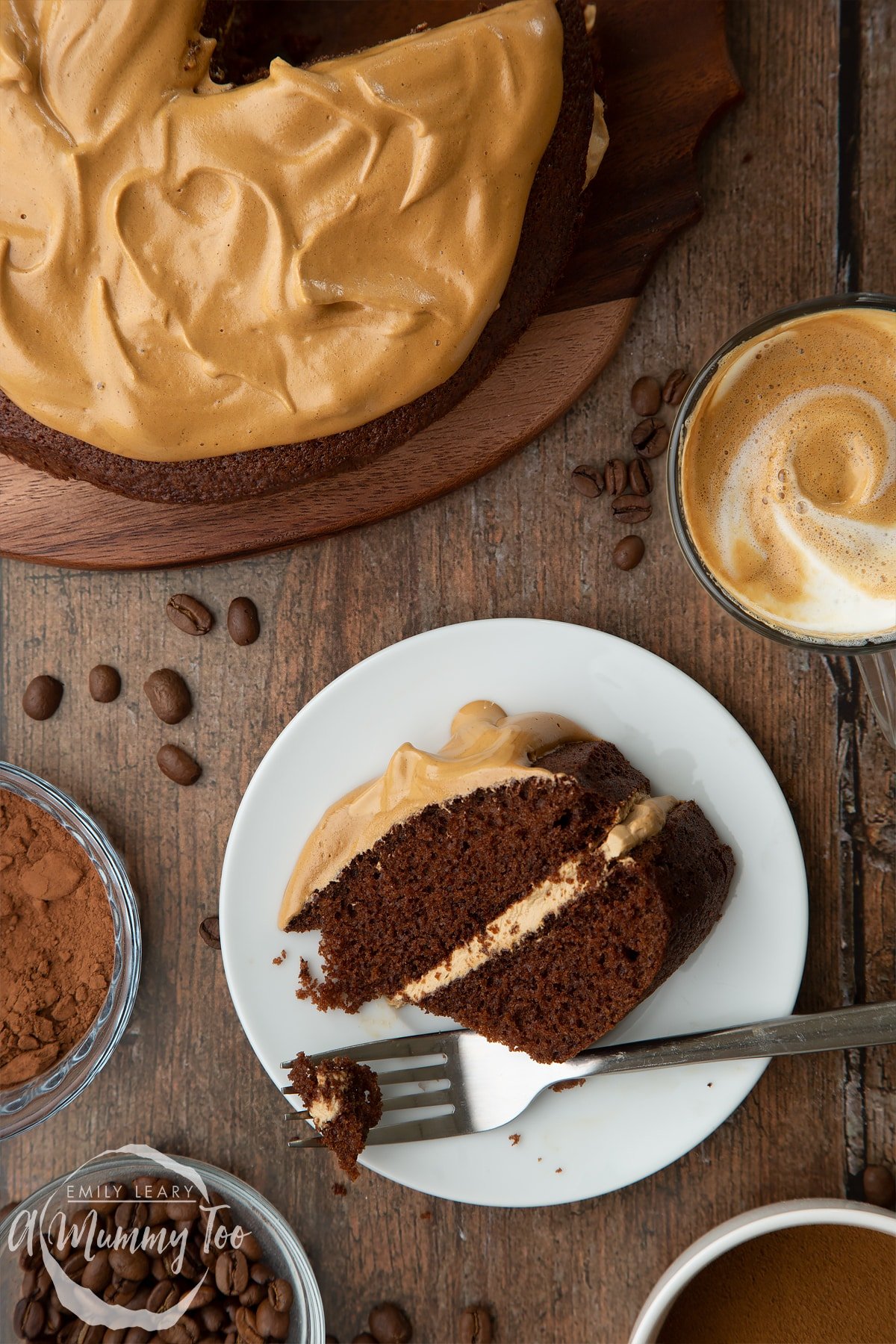 A slice of dalgona coffee cake on a white plate. A fork has cut into it. A cup of dalgona coffee, cocoa and coffee beans are shown to the sides.