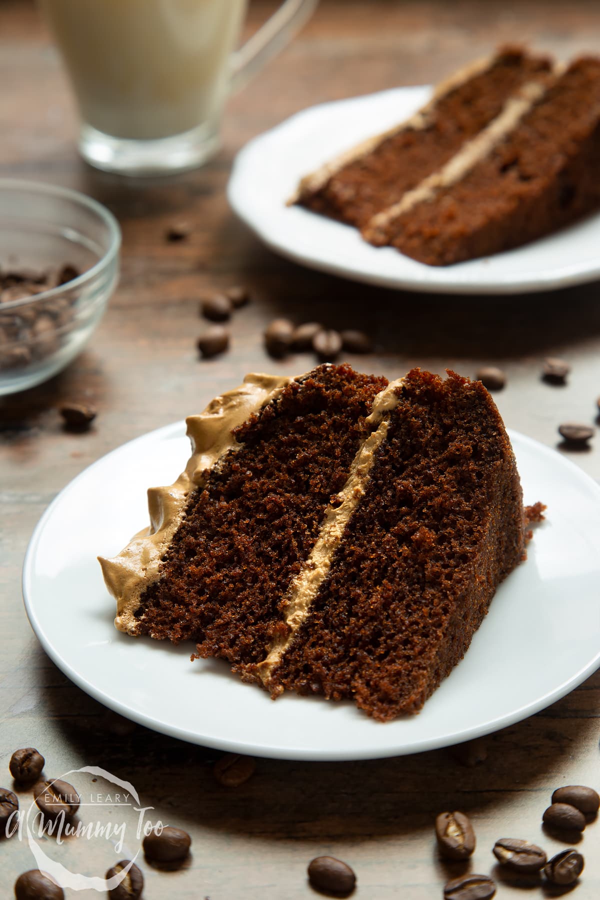 A slice of dalgona coffee cake on a white plate. A cup of dalgona coffee, coffee beans and another slice of cake is in the background.